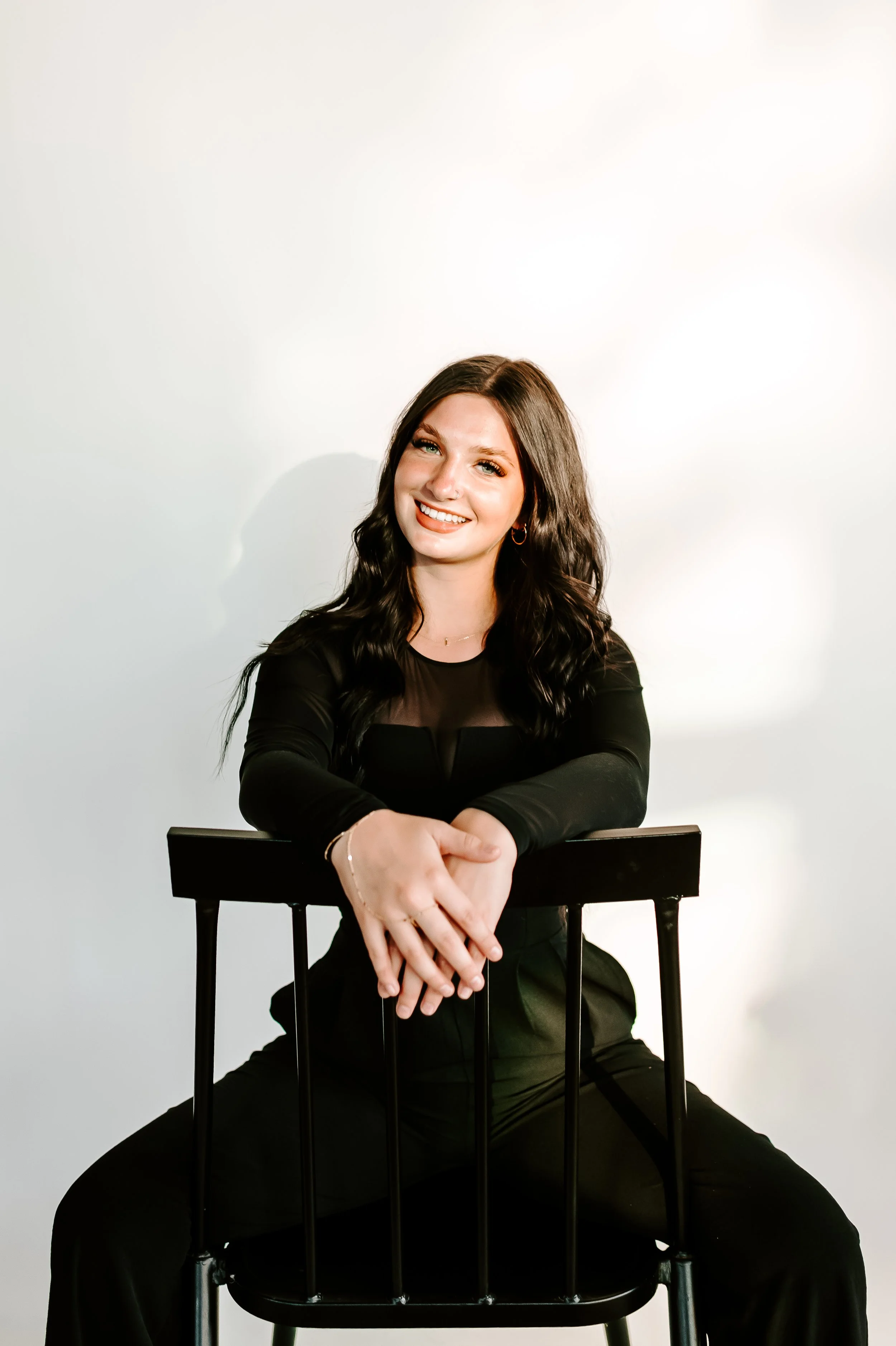 A young woman with long dark hair, smiling and sitting on a black chair against a plain white background, wearing black clothing.