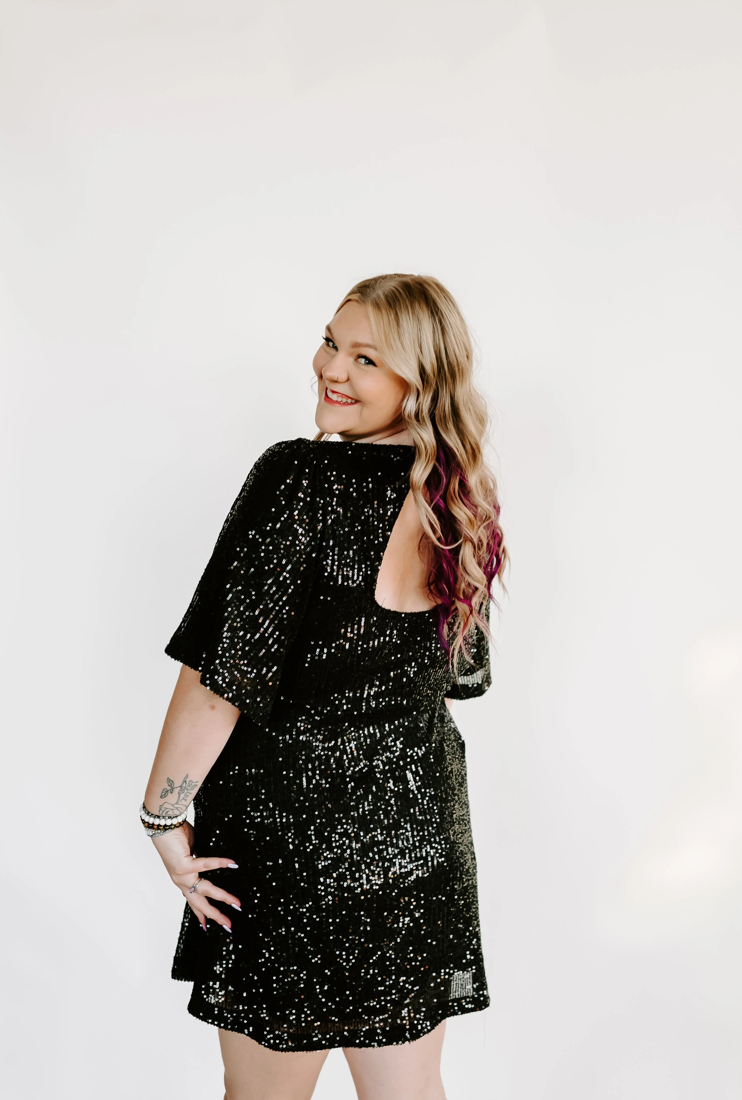 A woman with blonde, wavy hair with purple highlights, smiling and looking over her shoulder, wearing a black, sequined dress with an open back, standing against a plain white background.