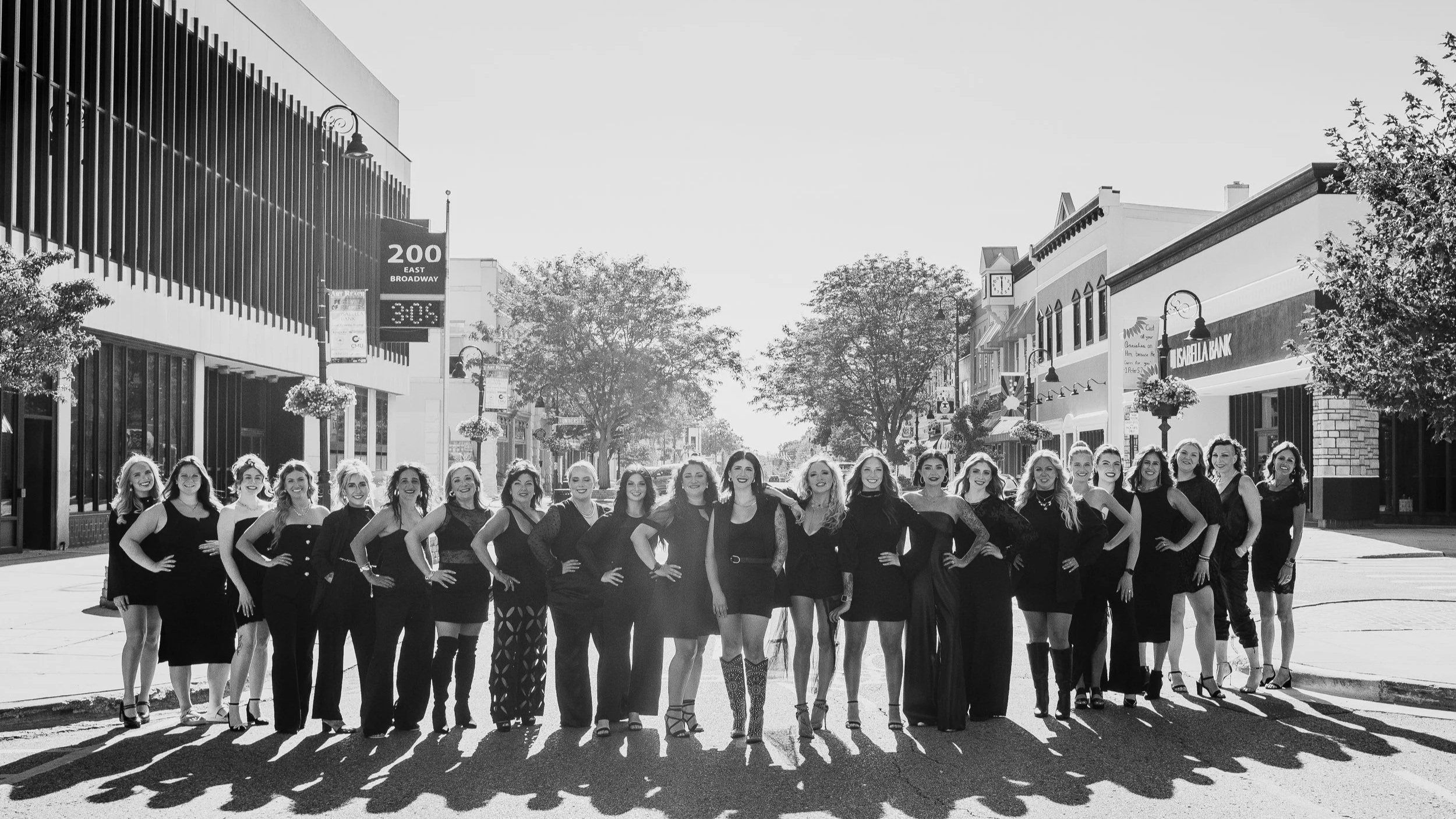 Group of women in stylish black outfits standing on a city street in sunlight, with shadows cast on the ground, trees, buildings, and street signs in the background.