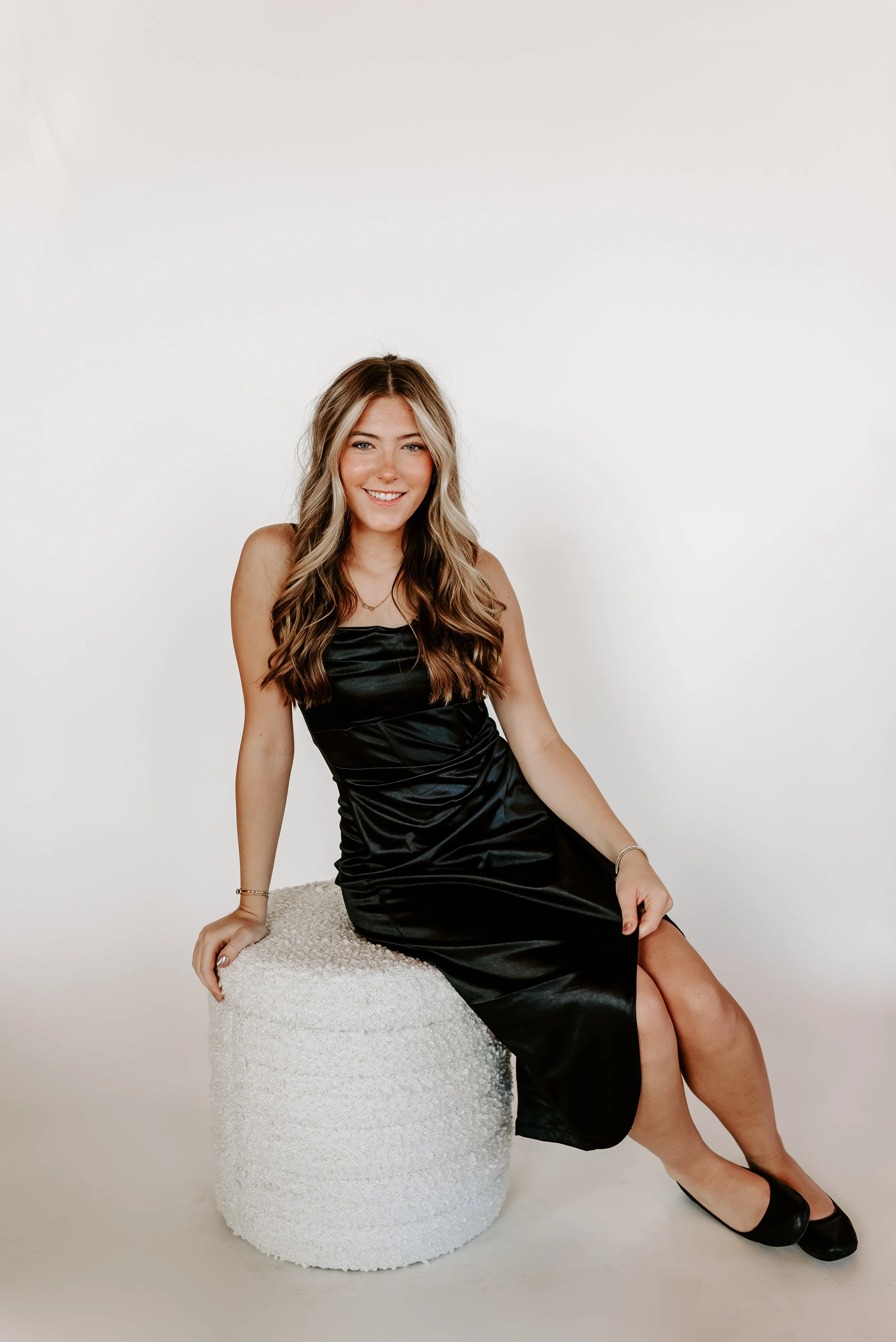 Young woman with long wavy hair, wearing a black satin dress, sitting on a textured white stool, smiling at the camera against a plain white background.