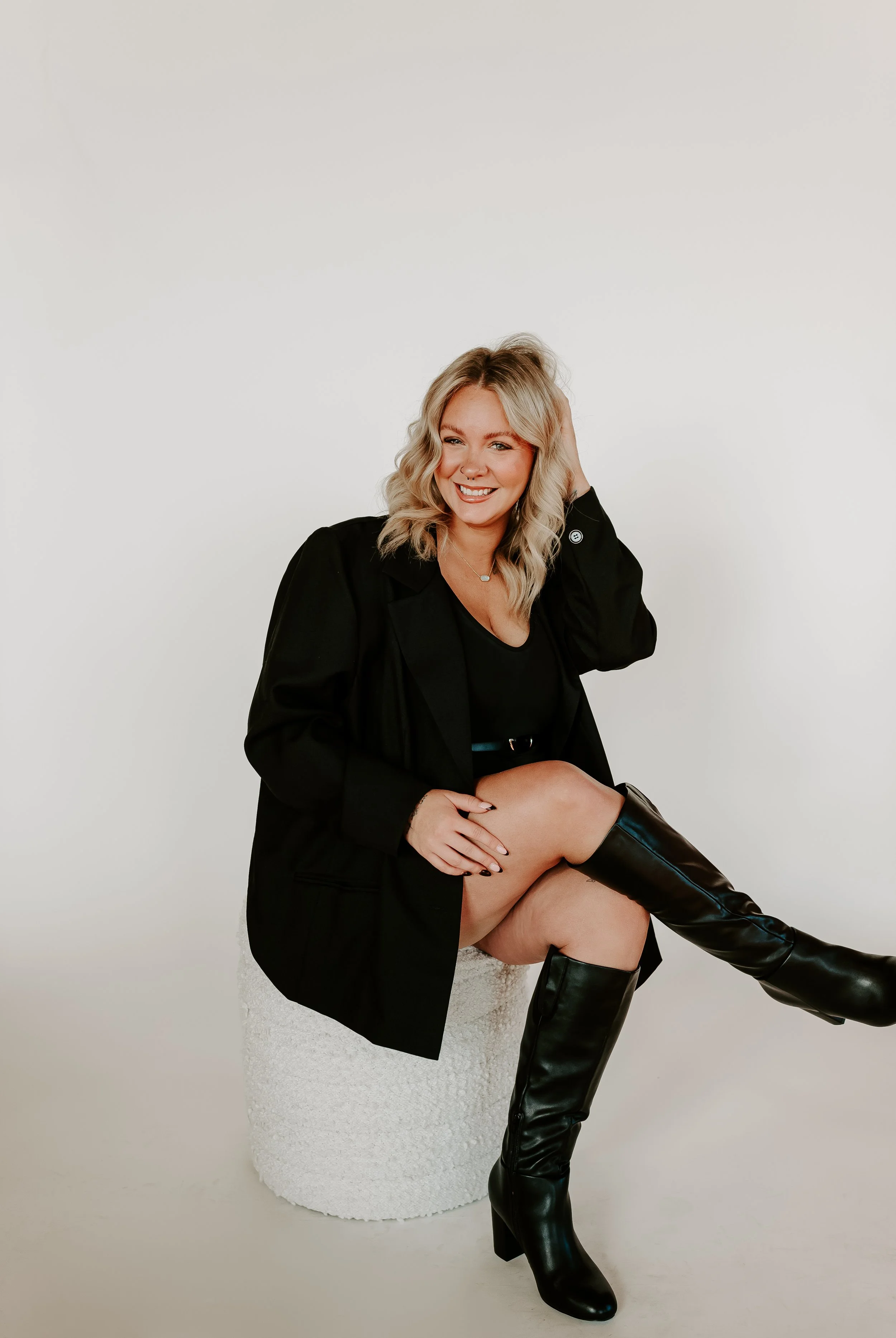 A woman with blonde wavy hair, wearing a black blazer, black top, and black over-the-knee boots, sitting on a white textured stool, smiling against a plain white background.