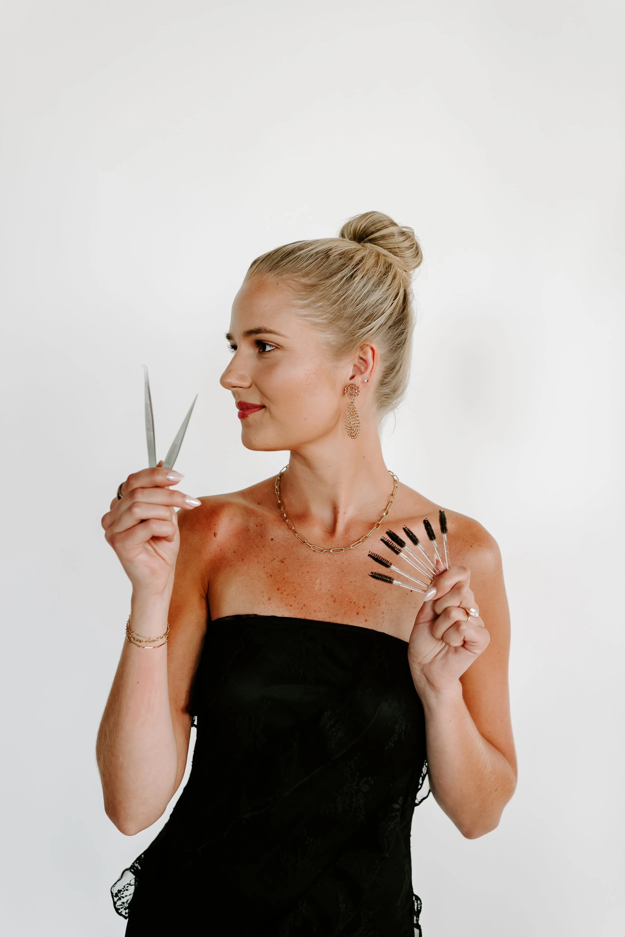 A woman with blonde hair in a bun, wearing a black strapless dress, holding hairdressing scissors and a set of makeup brushes, standing against a plain white background.