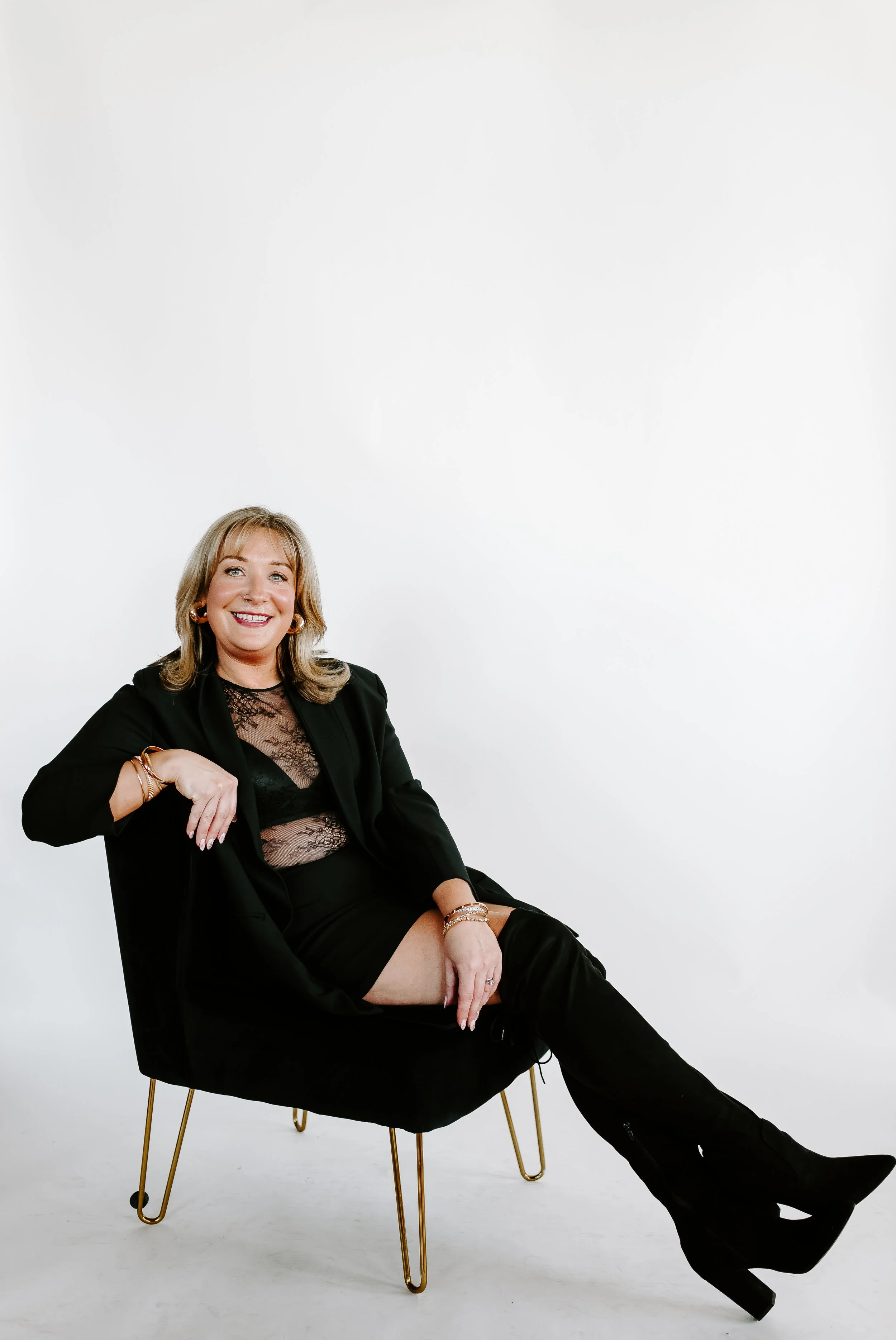 A woman sitting in a black chair with gold legs, smiling, wearing a black blazer, a lace top, a black skirt, and black thigh-high boots, against a plain white background.