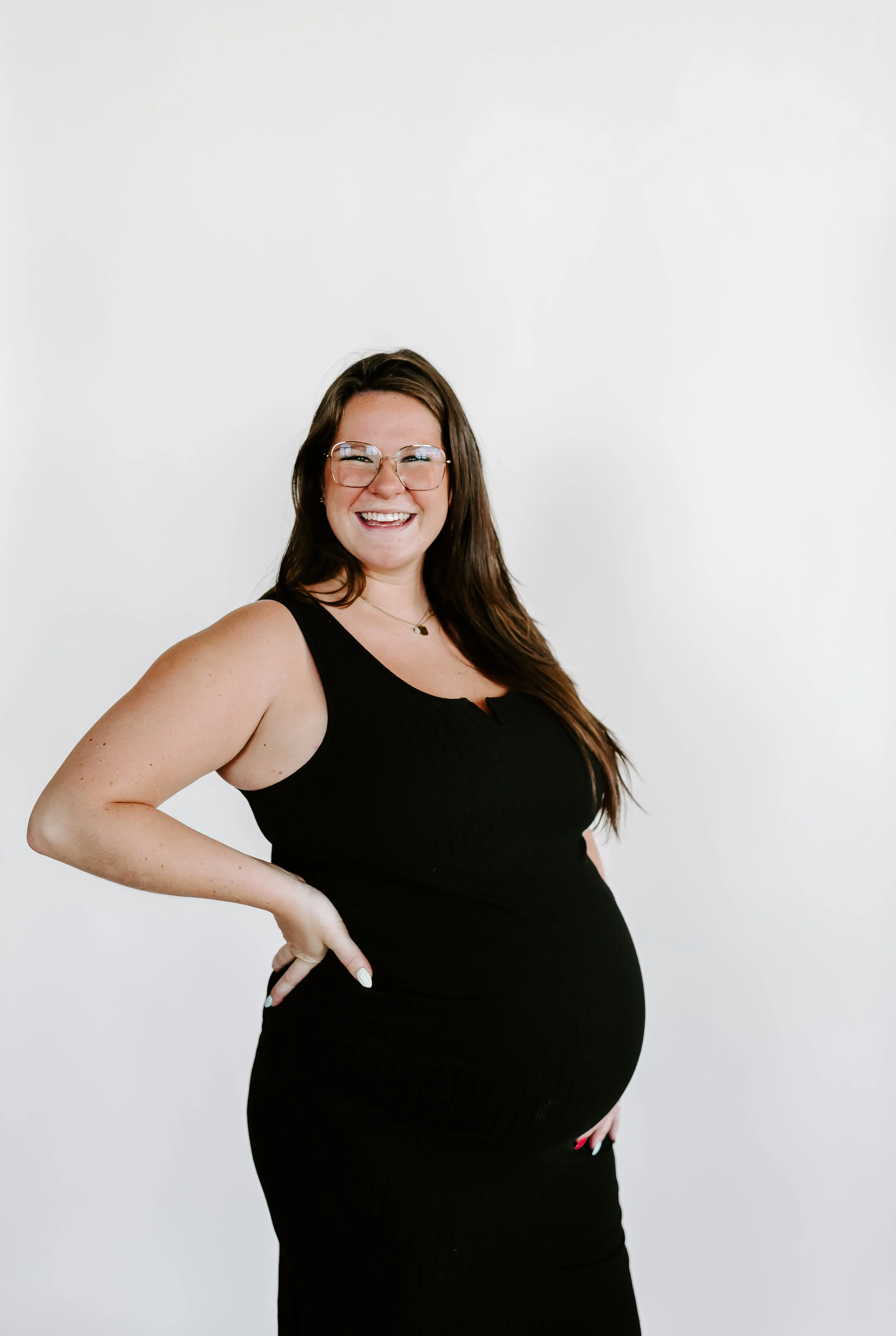 A pregnant woman with long brown hair, glasses, and a wide smile, wearing a sleeveless black dress, standing against a plain white background.