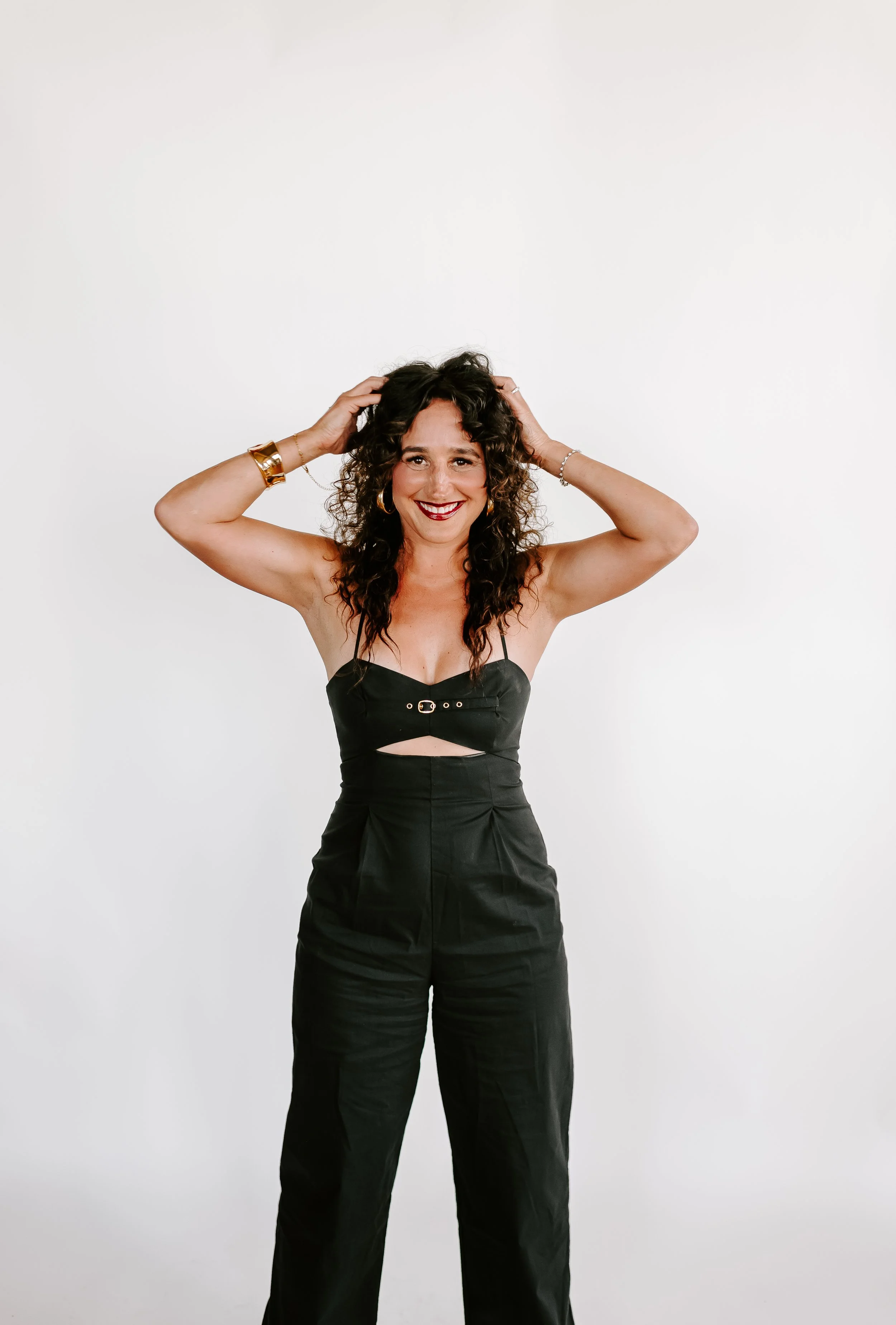 Woman with curly dark hair wearing a black outfit posing with hands in her hair against a plain white background.