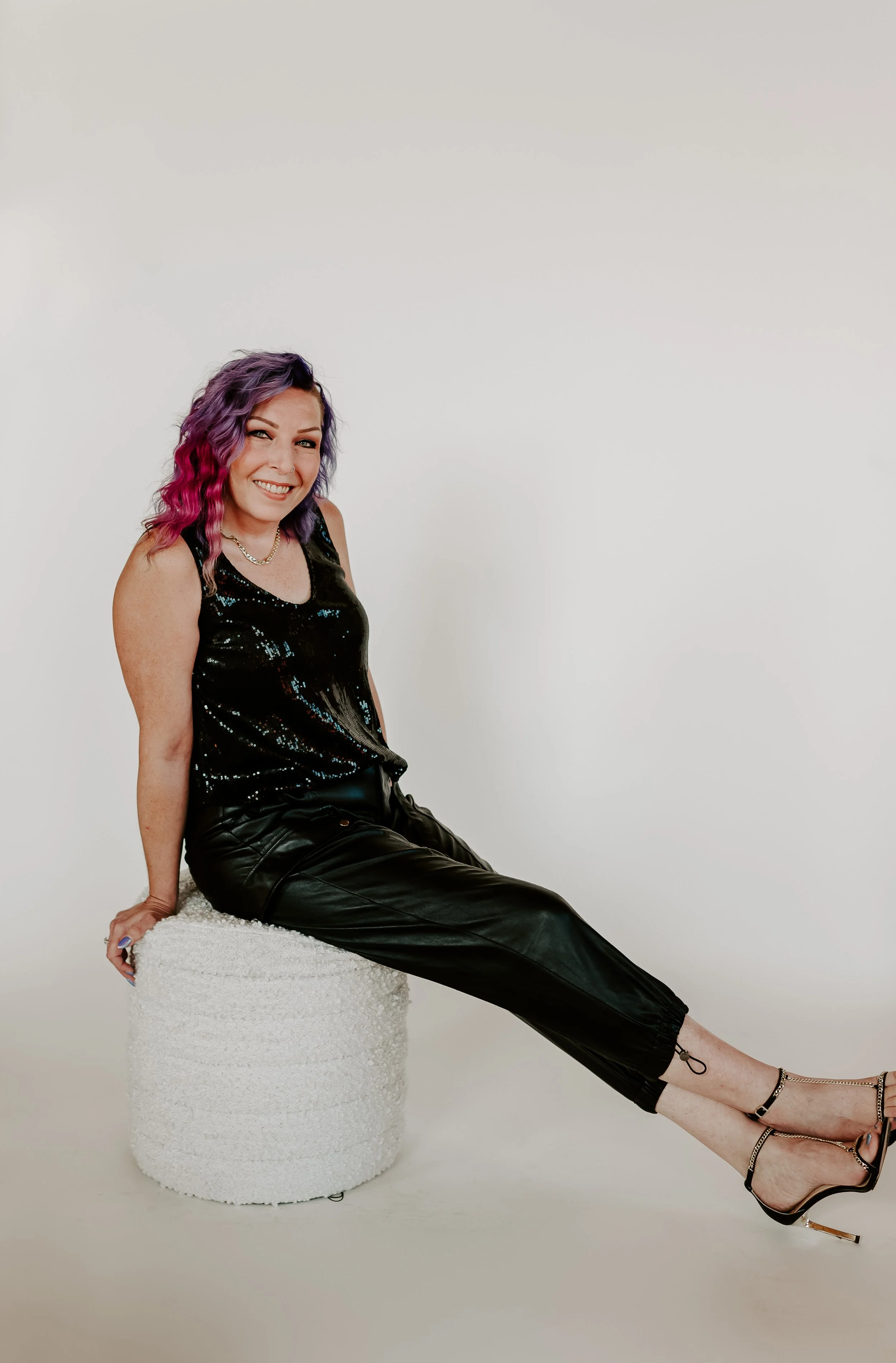 A woman with purple and pink hair sitting on a white textured stool against a plain white background, wearing a black sequined top, black leather pants, and high-heeled sandals, smiling at the camera.