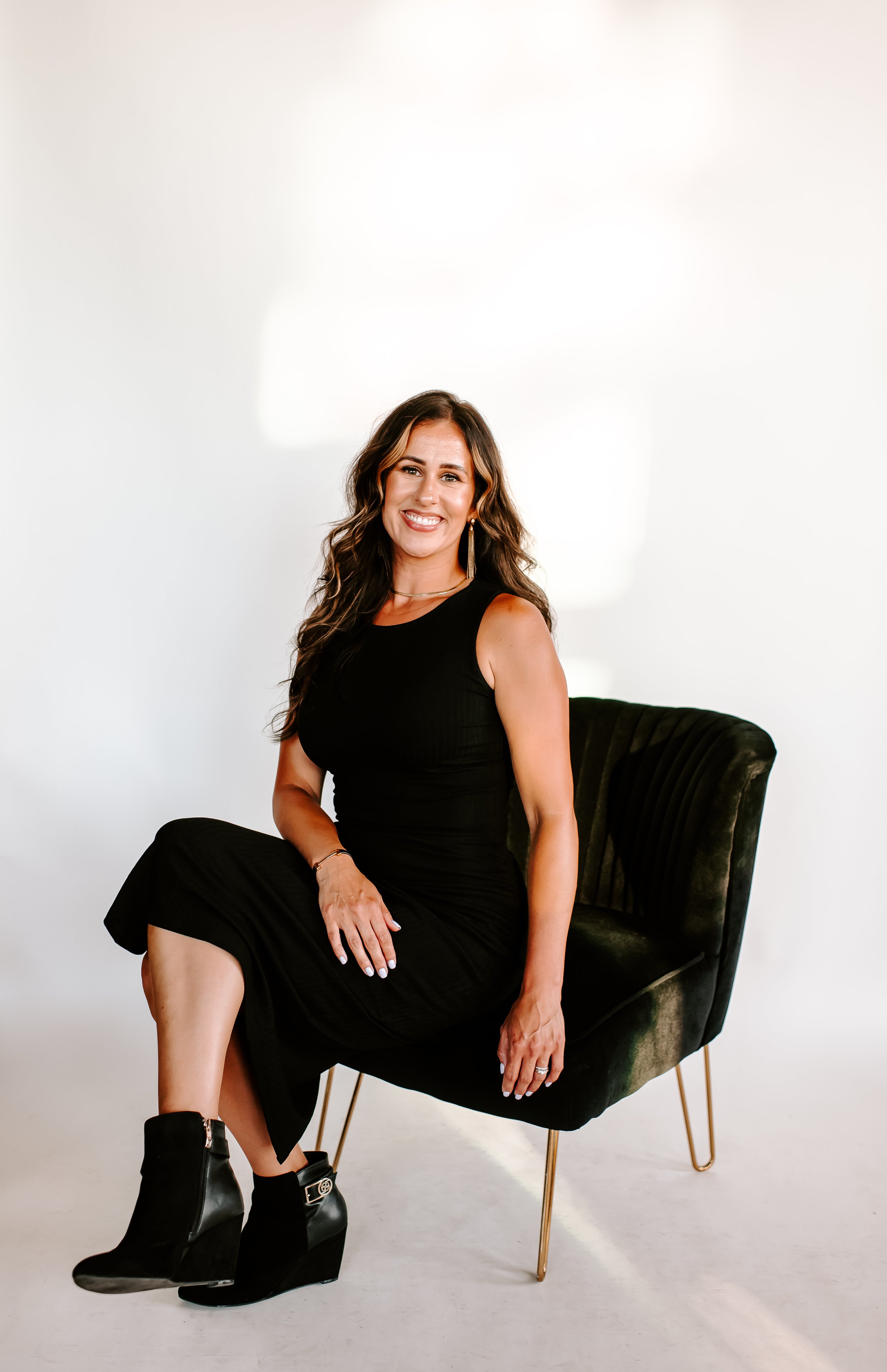 A woman sitting on a black velvet chair with gold legs, smiling, wearing a sleeveless black dress and black wedge ankle boots, against a plain white background.