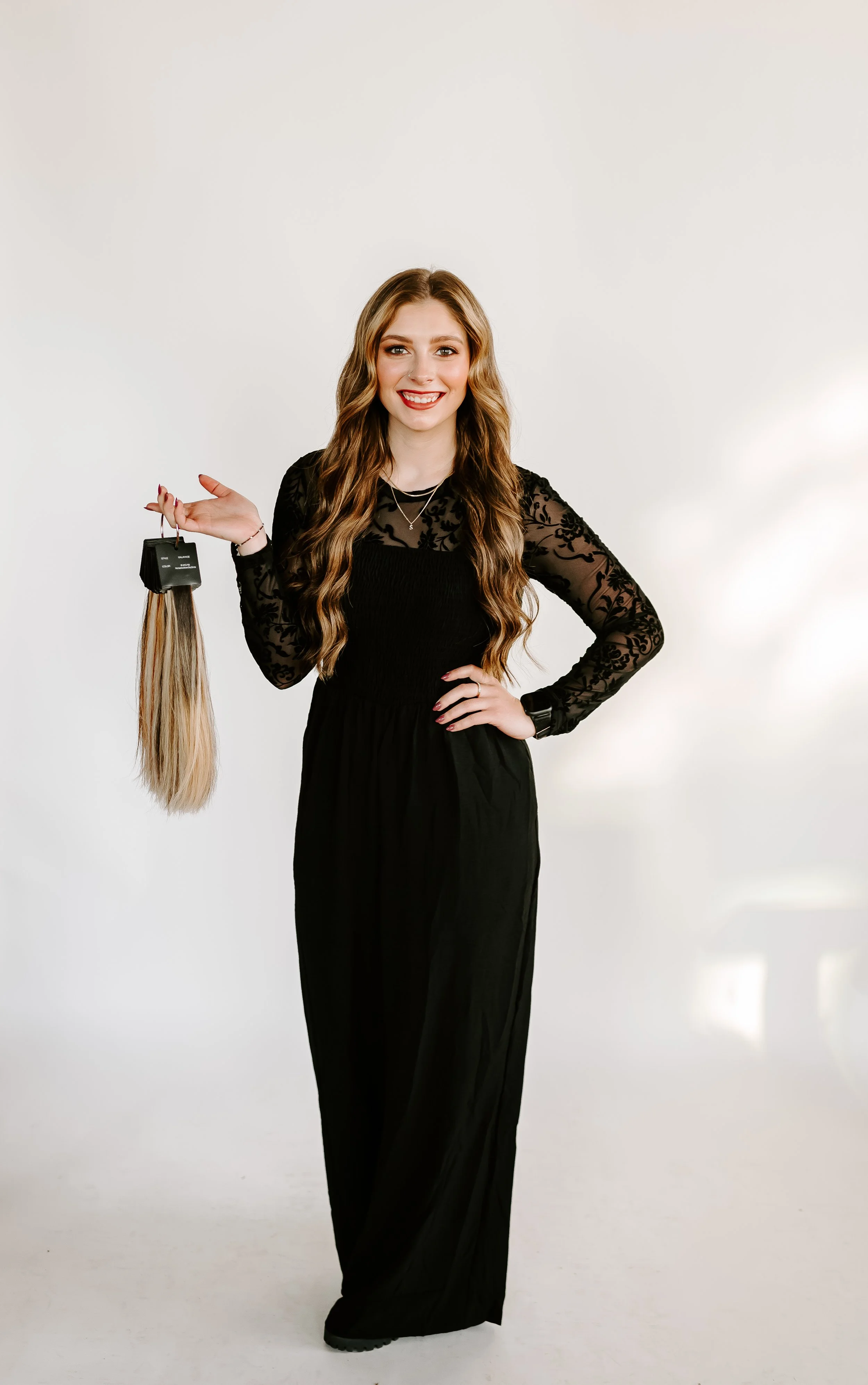 Woman with long wavy hair wearing a black dress, smiling, holding a hair extension in her right hand, standing against a plain white background.