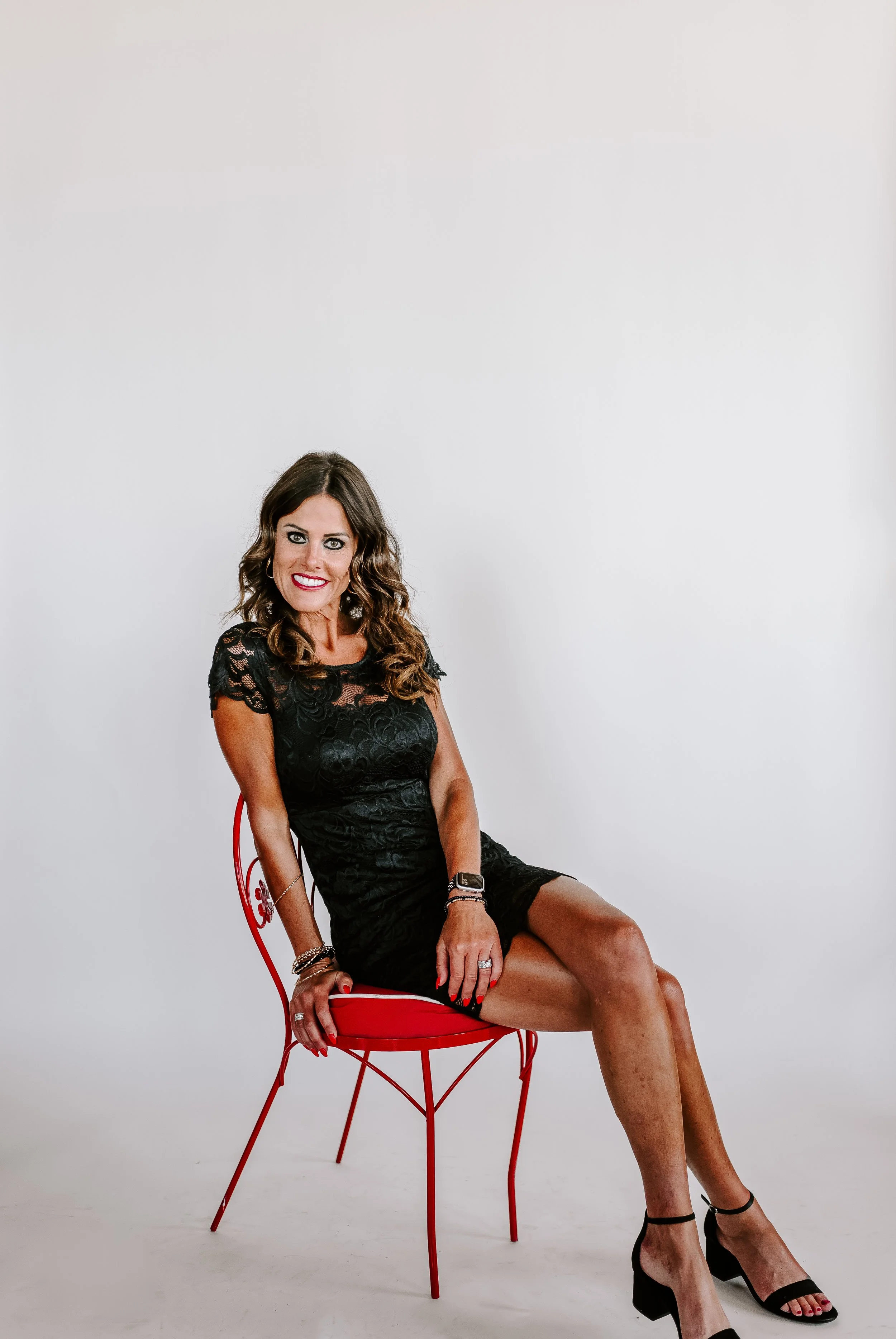 Woman with brown hair wearing a black lace dress, sitting on a red chair against a plain white background, smiling at the camera.