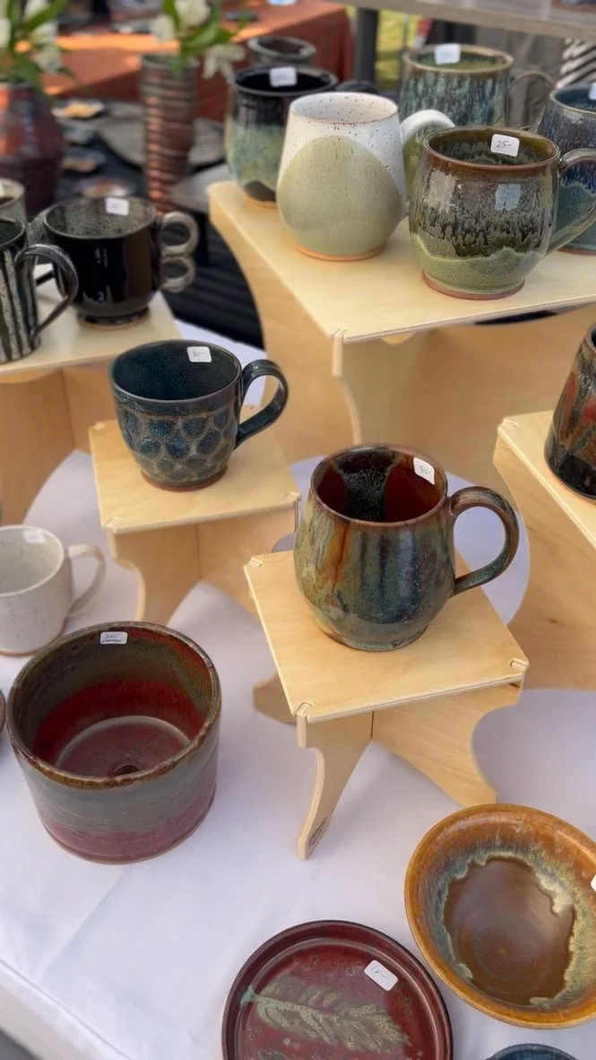 Various handmade ceramic mugs and bowls displayed on wooden stands and tables at a craft fair.