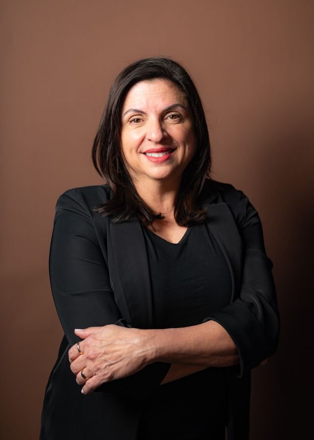 A woman with shoulder-length dark hair smiling, wearing a black blazer and a black top, standing against a plain brown background.