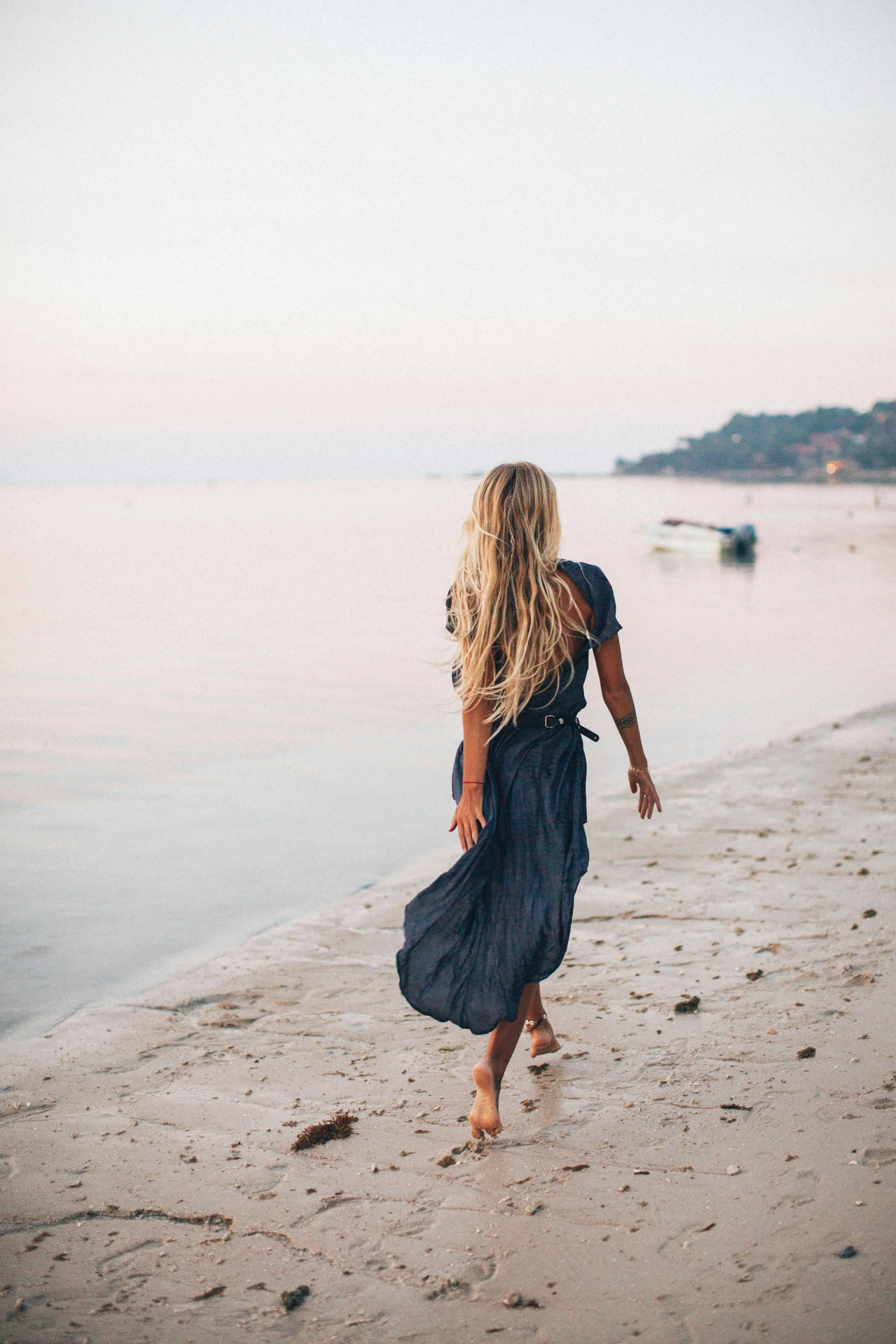 Woman with long blonde hair in a blue dress walking barefoot on the beach during sunset