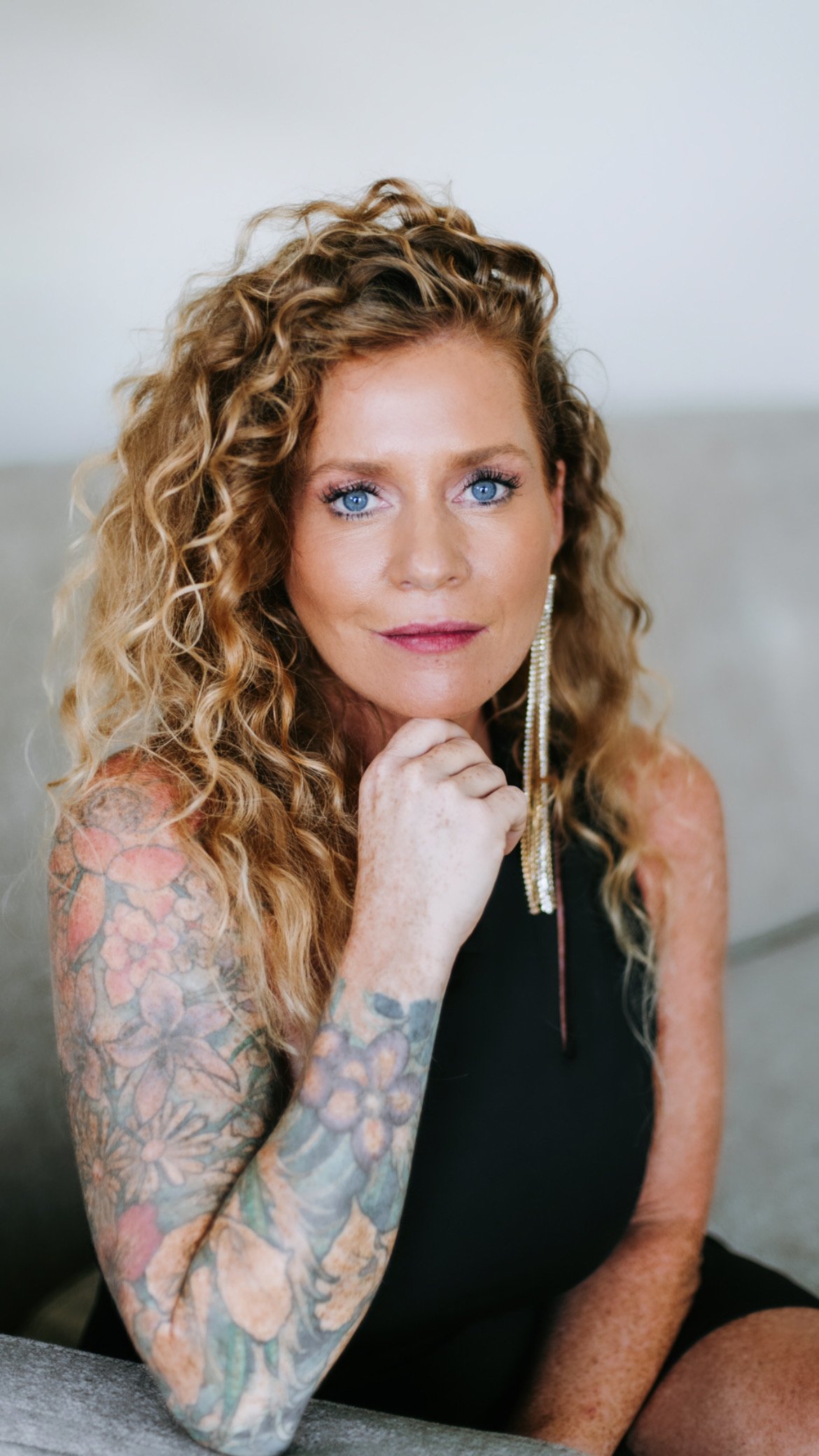 A woman with curly red hair, blue eyes, and tattoos on her arm, wearing a black top and long earrings, sitting on a sofa.