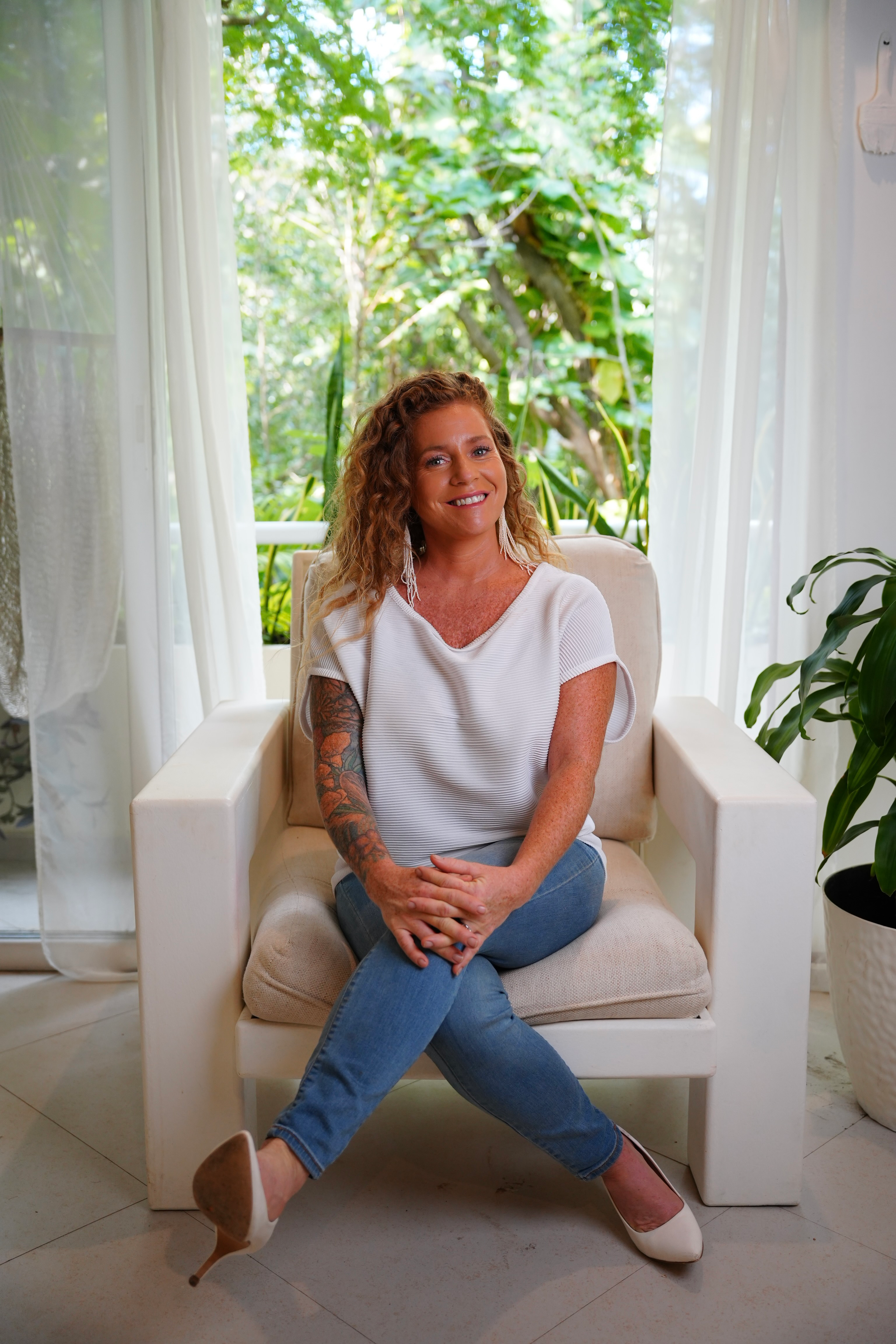 A woman with curly hair, tattoos on her left arm, wearing a white blouse and blue jeans, sitting on a white armchair with her legs crossed in a bright room with large windows and green plants outside.