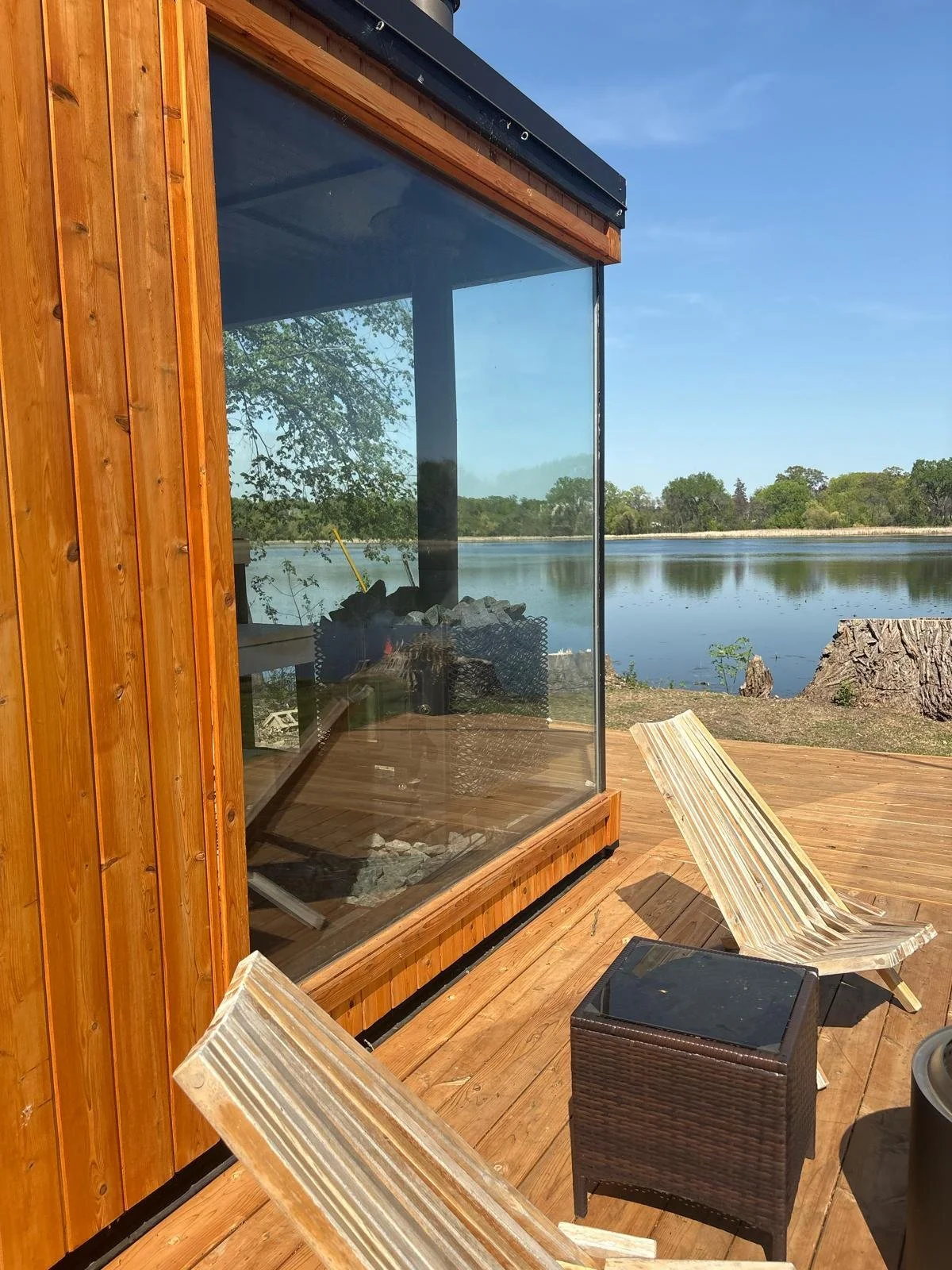 A lakeside wooden deck with two wooden lounge chairs, a small wicker table, and a glass-walled structure reflecting the trees and sky, overlooking a calm lake with trees in the background on a clear, sunny day.