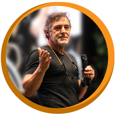 A man with gray hair and a beard, wearing a black shirt and glasses hanging around his neck, holding a microphone, gesturing with his right hand.