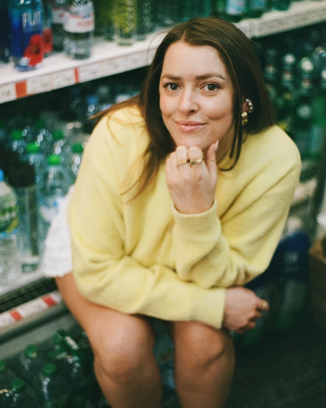 A woman with long brown hair, wearing a yellow sweater, sitting in a grocery store aisle surrounded by soda bottles, smiling at the camera, resting her chin on her hand.