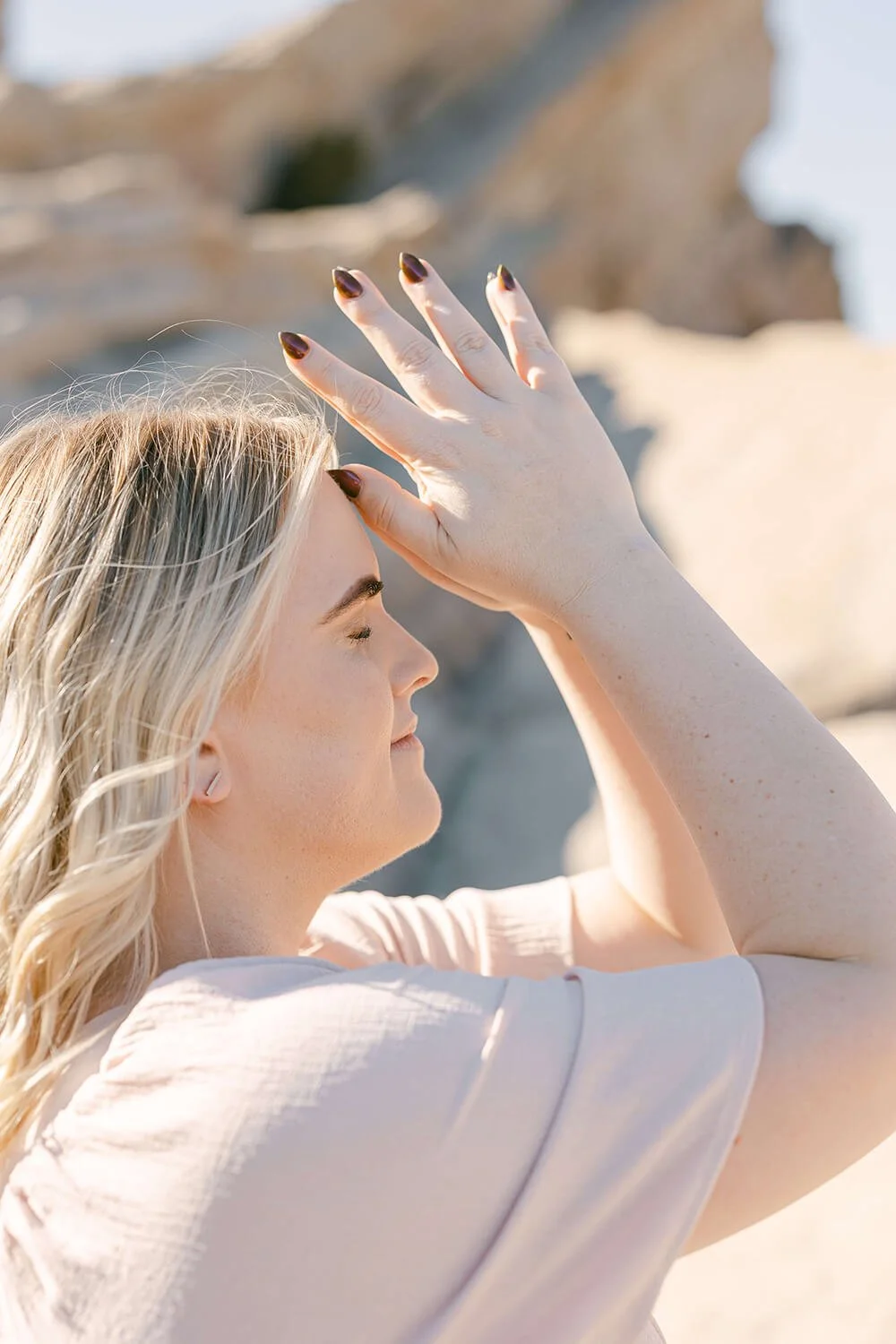 A woman with blond hair and dark nail polish is closing her eyes with her hand raised, touching her forehead, outdoors in a sunny location with rocky terrain in the background.