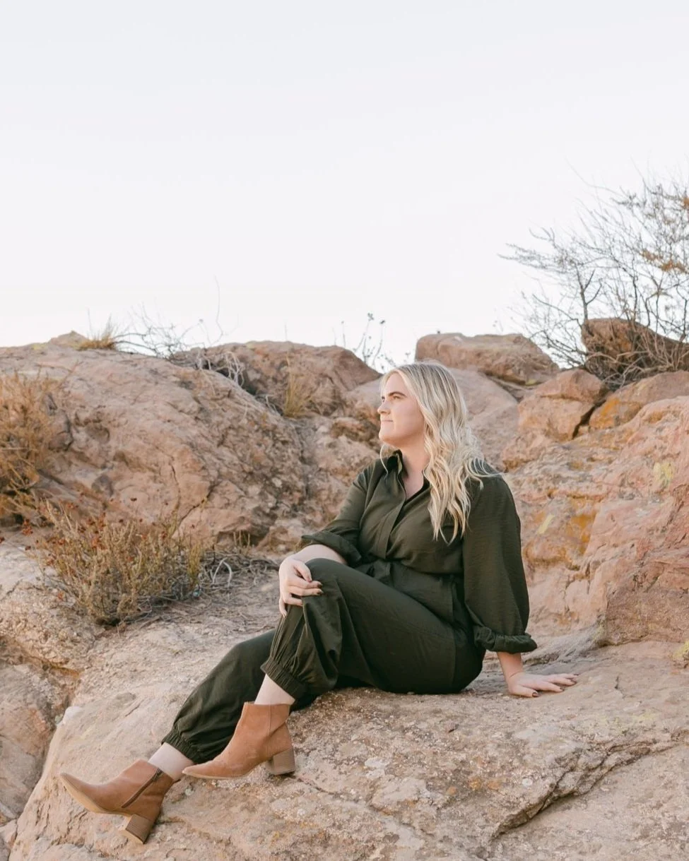 A woman with blonde hair sitting on a large rock in a desert landscape, wearing a dark green outfit and tan ankle boots, looking into the distance.