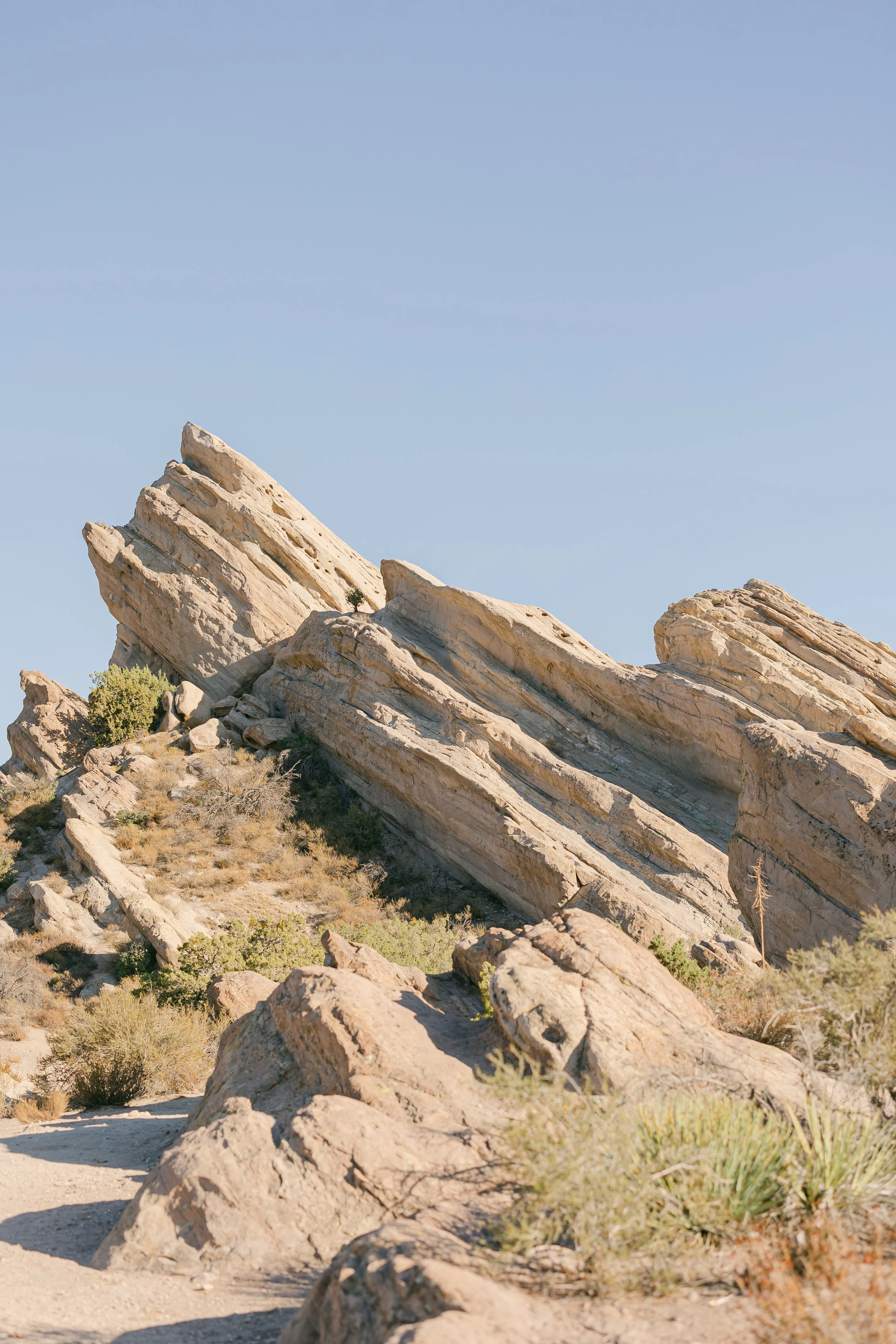 Large tilted rock formations in a desert landscape with sparse vegetation and a clear blue sky.