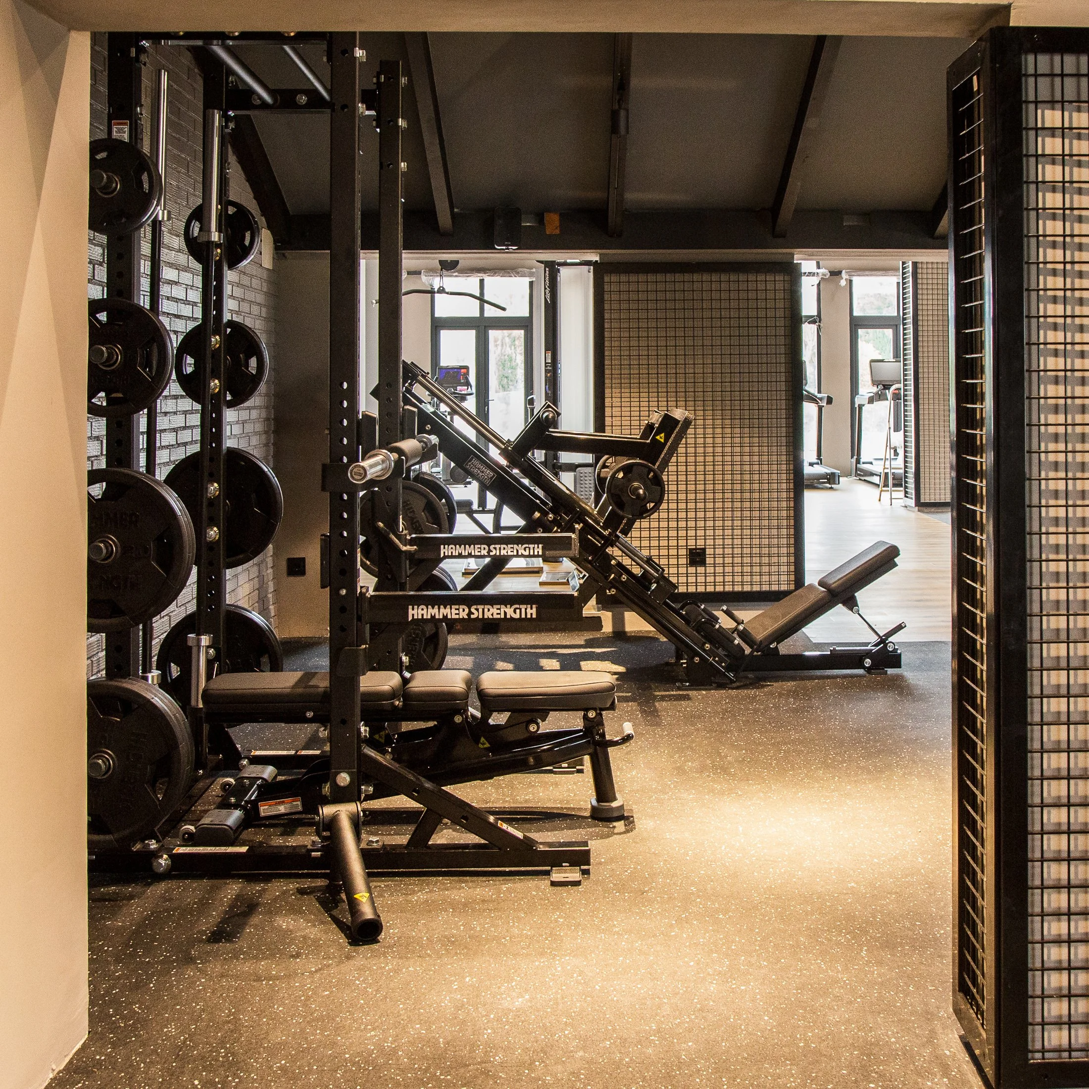 A modern gym with weightlifting equipment, including a squat rack with weights, an adjustable bench, and a leg press machine, with large windows providing natural light.