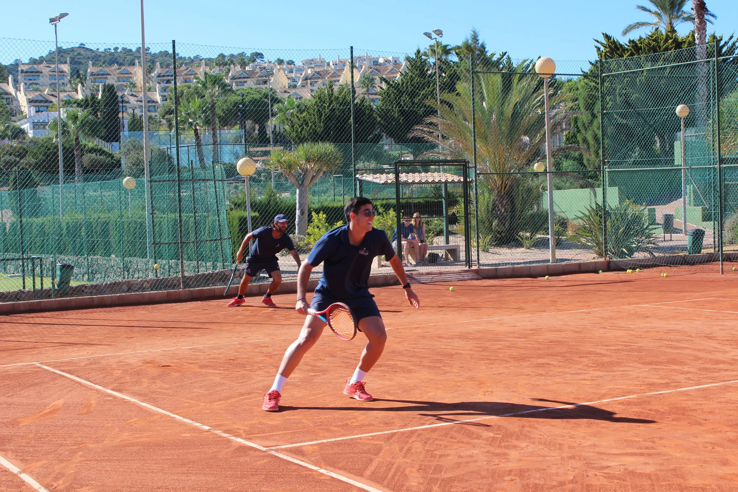 A tennis coach practicing with a team on a clay tennis court. The coach is holding tennis balls and a racket, with a cart filled with tennis balls beside her. Four players are on the court, preparing for practice or a game, with a backdrop of trees and white buildings.