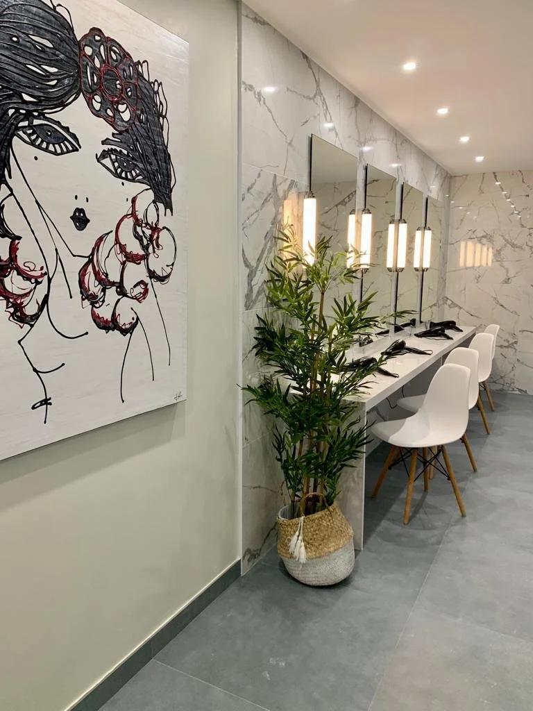 Modern salon with white chairs, marble wall, large mirror, potted plant, and artwork of a woman with flowers in her hair.