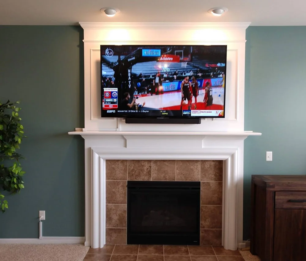 Living room fireplace with a mounted flat-screen TV showing a basketball game, a potted plant on the left, and a wooden cabinet on the right.