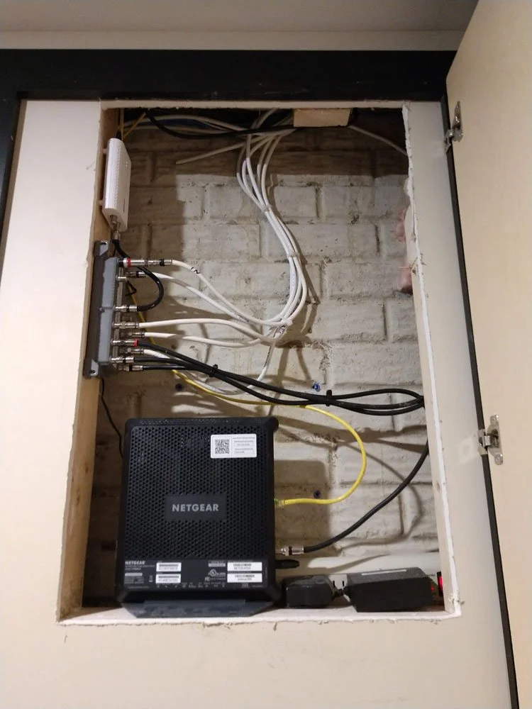 Open wall cabinet with network equipment, including a NETGEAR modem/router, connected cables, and a splitter with multiple connections.