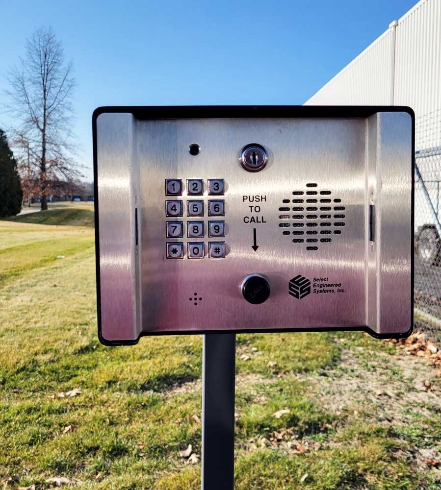 An outdoor metallic intercom or calling device with a keypad, a speaker, and a keyhole, mounted on a pole in a grassy area with trees and clear blue sky in the background.