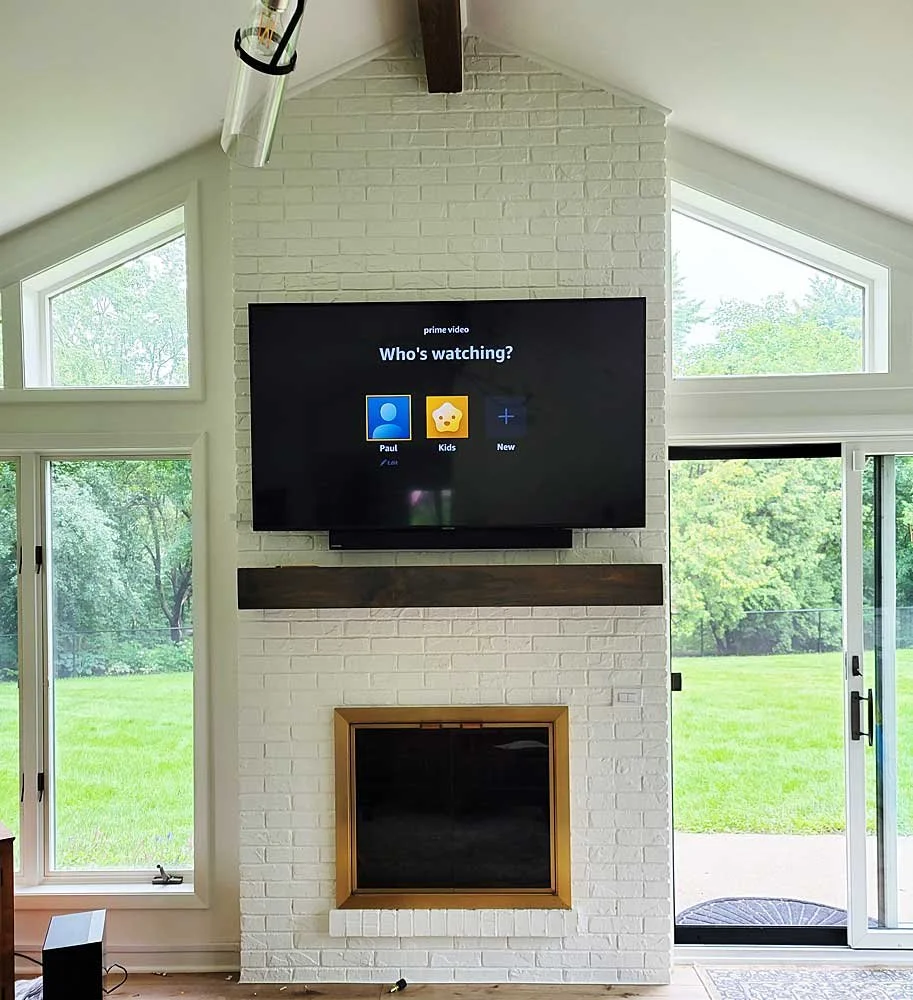 Living room with a white brick fireplace, a wall-mounted TV showing the user profile for Prime Video, large windows, and a sliding glass door leading to a grassy outdoor area.