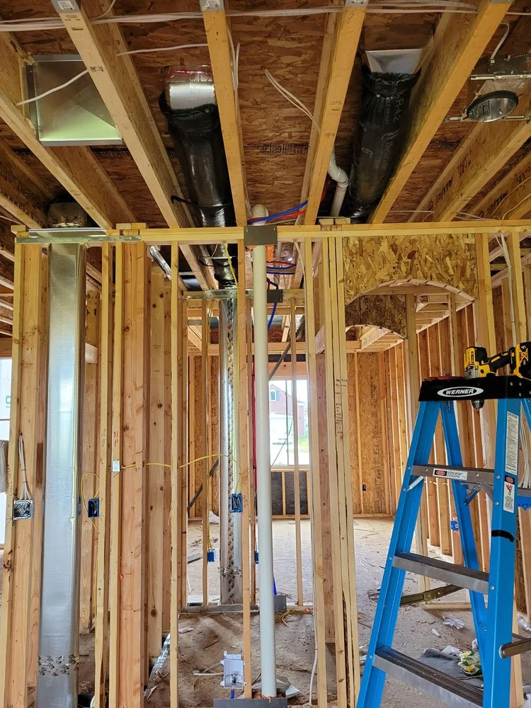 Interior view of a house under construction with exposed wooden framing, ventilation ducts, electrical wiring, and a blue step ladder.