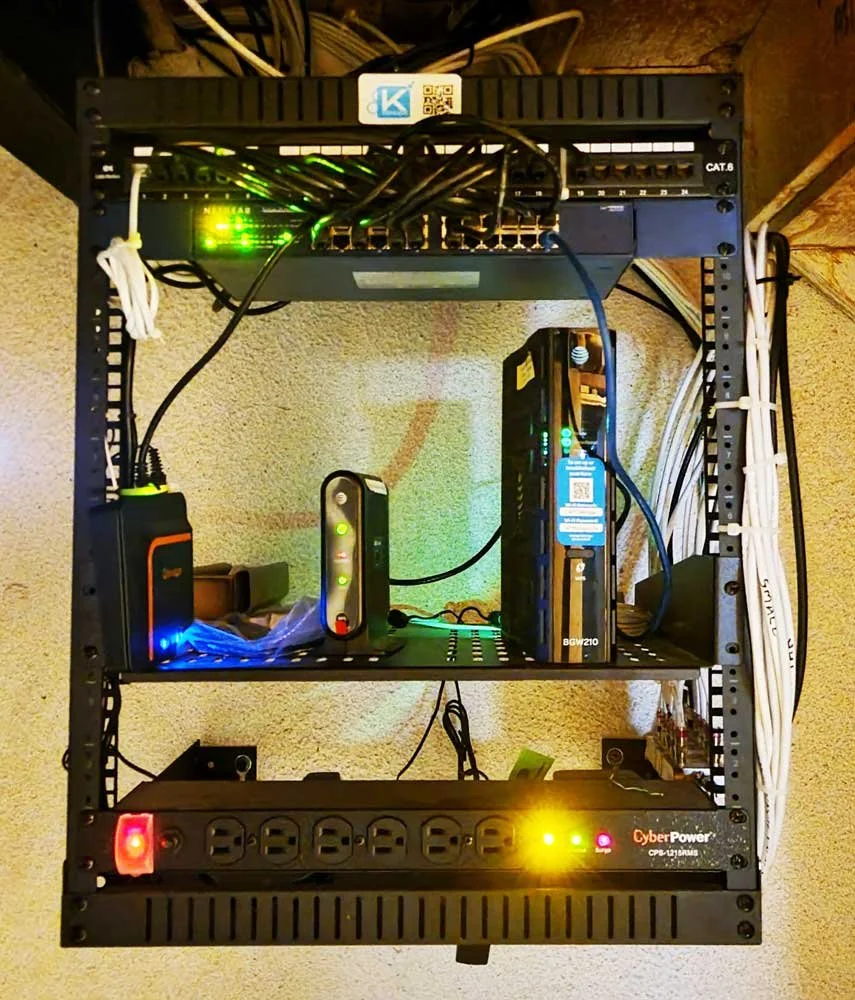 Network server rack with various electronic devices, power strips, and cables installed on black metal shelves against a beige wall.