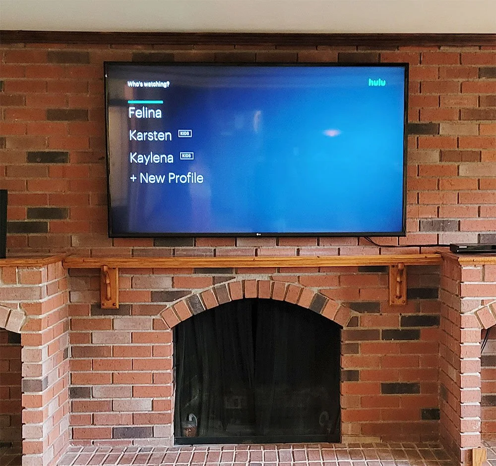 TV screen displaying a streaming service's Watchlist with four profiles named Fellina, Karsten, Kaylena, and an option to add a new profile, mounted above a brick fireplace with a wooden mantel.