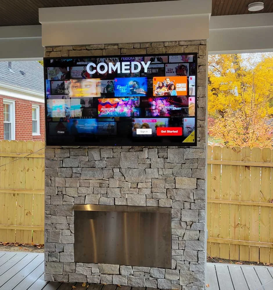 TV screen displaying streaming service home page with the word 'COMEDY' at the top, mounted on a stone pillar outside with a wooden fence and trees with yellow leaves in the background.
