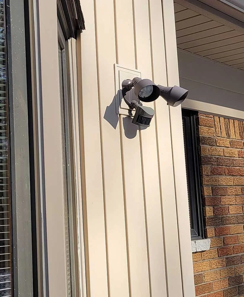 Exterior wall of a house with beige siding, a window with blinds, and a security camera mounted on the wall.
