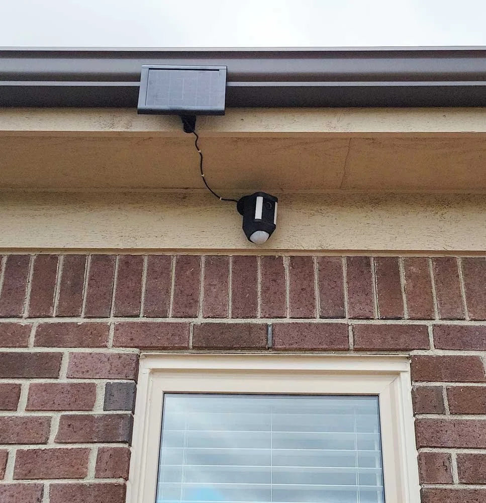 Exterior view of a house showing a brick wall, window with white frame and blinds, and a security camera mounted on the wall above the window. A solar panel is attached nearby with a cable connected to the camera.