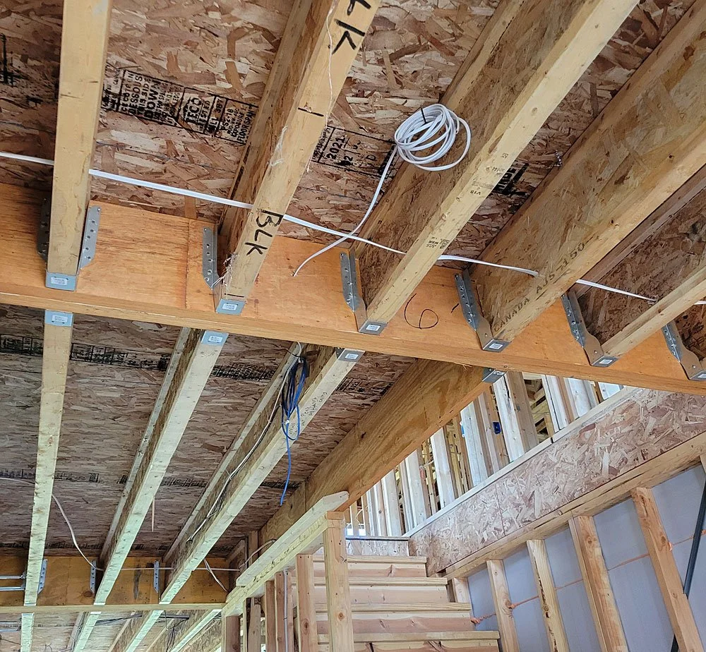 Interior view of a building under construction with exposed wooden studs, beams, wiring, and plywood sheathing on the ceiling and walls.