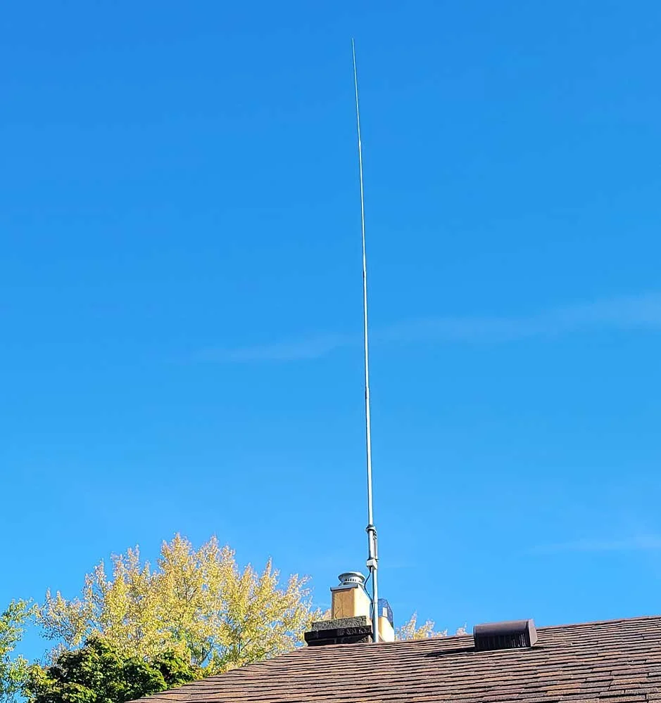 A tall radio or TV antenna mounted on a roof with a clear blue sky in the background and trees with autumn leaves.