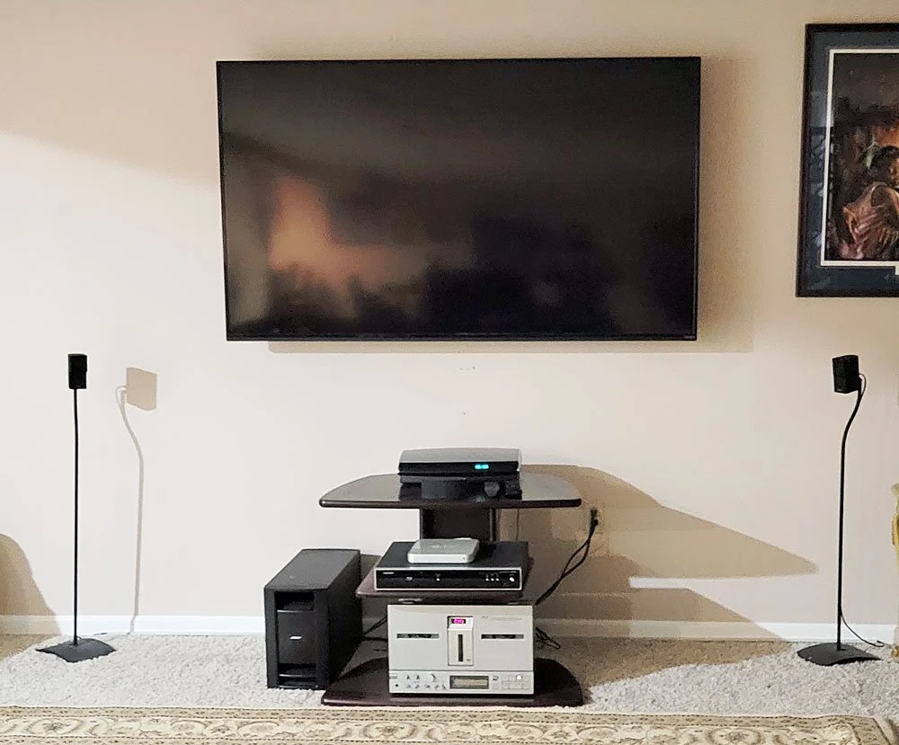 Living room entertainment setup with a large flat-screen TV mounted on the wall, a media stand with electronic devices, and two tall black speakers on the floor.