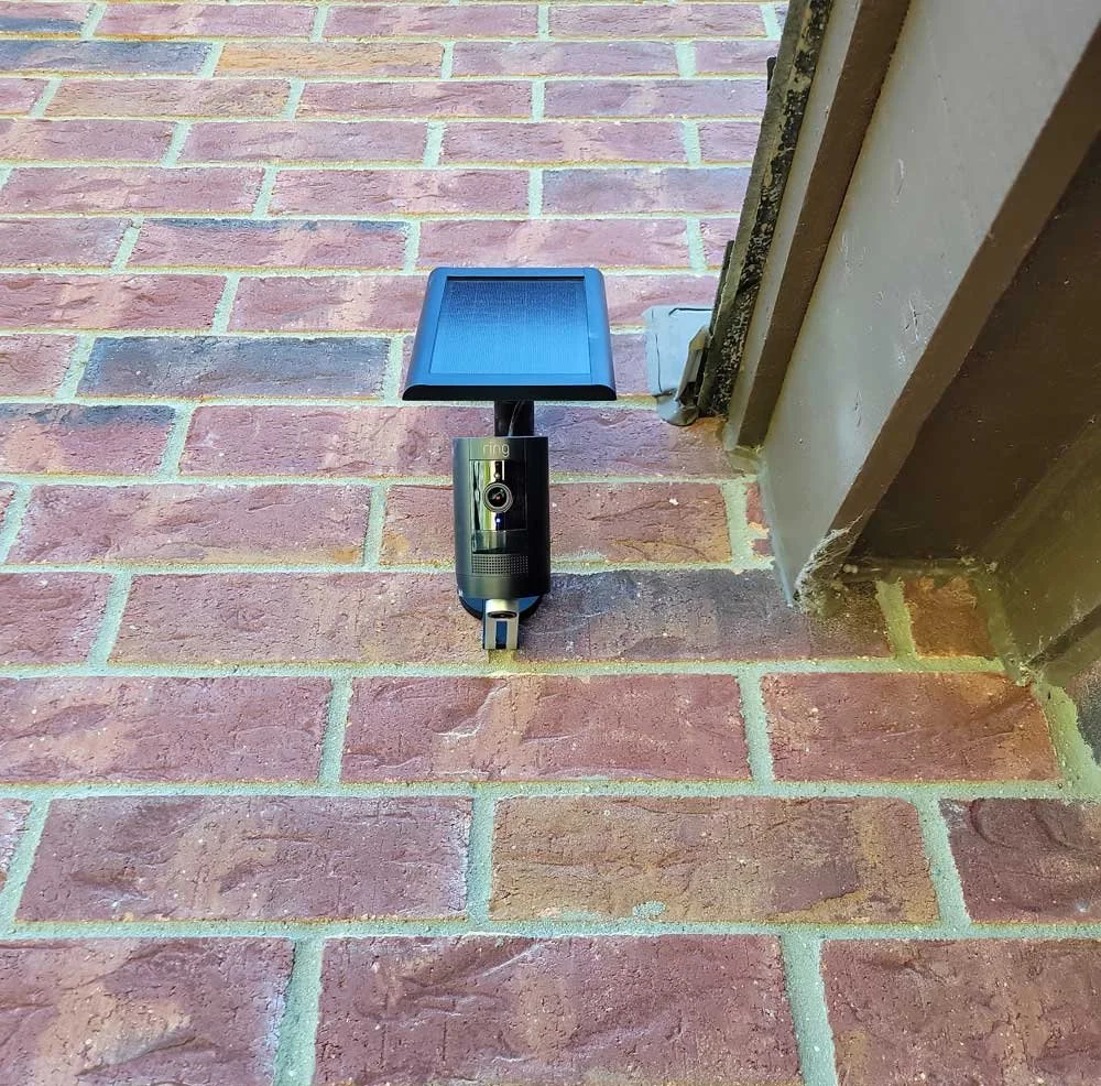 A Ring security camera with a solar panel mounted on a brick porch next to a door frame.