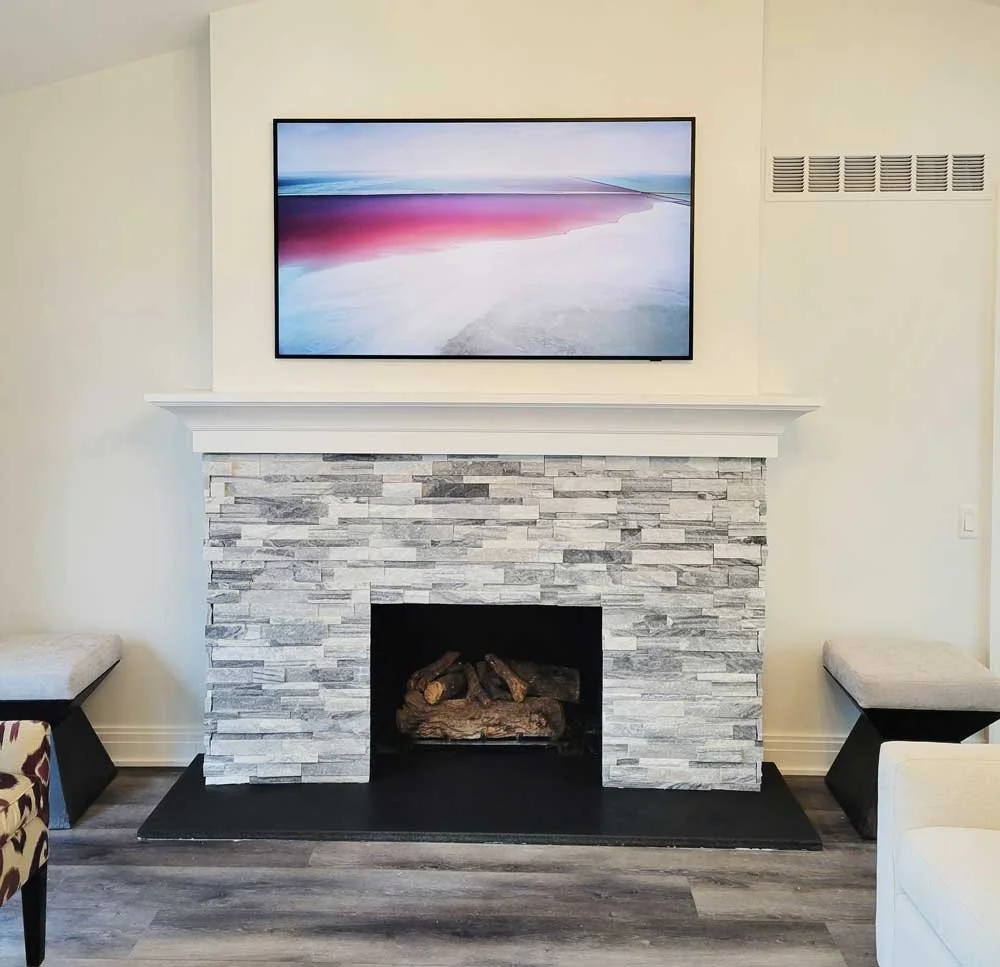 Living room with a gray stacked stone fireplace, white mantel, and a flat-screen TV mounted above. Two benches are on either side of the fireplace, and part of a patterned chair is visible in the bottom left corner.