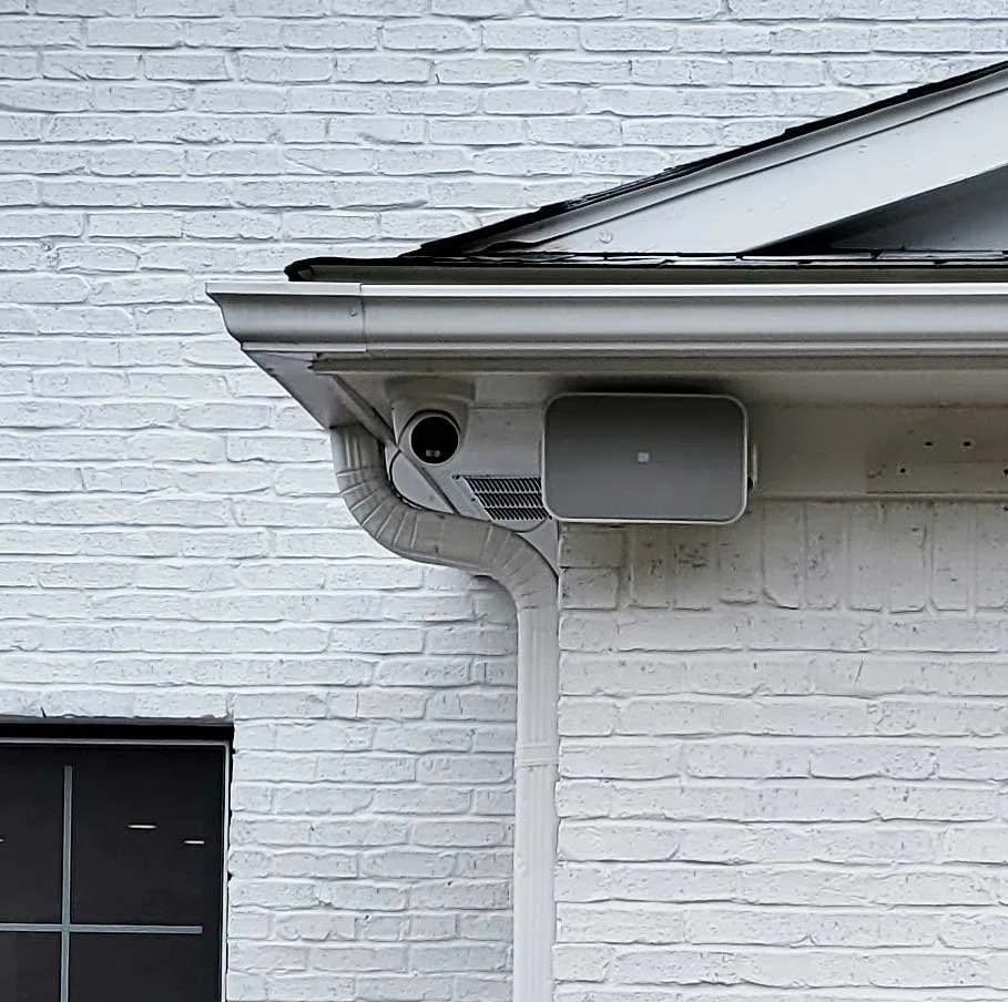 Close-up of the gutter and brick wall of a house, showing a security camera, a yellow box, and a white downspout.
