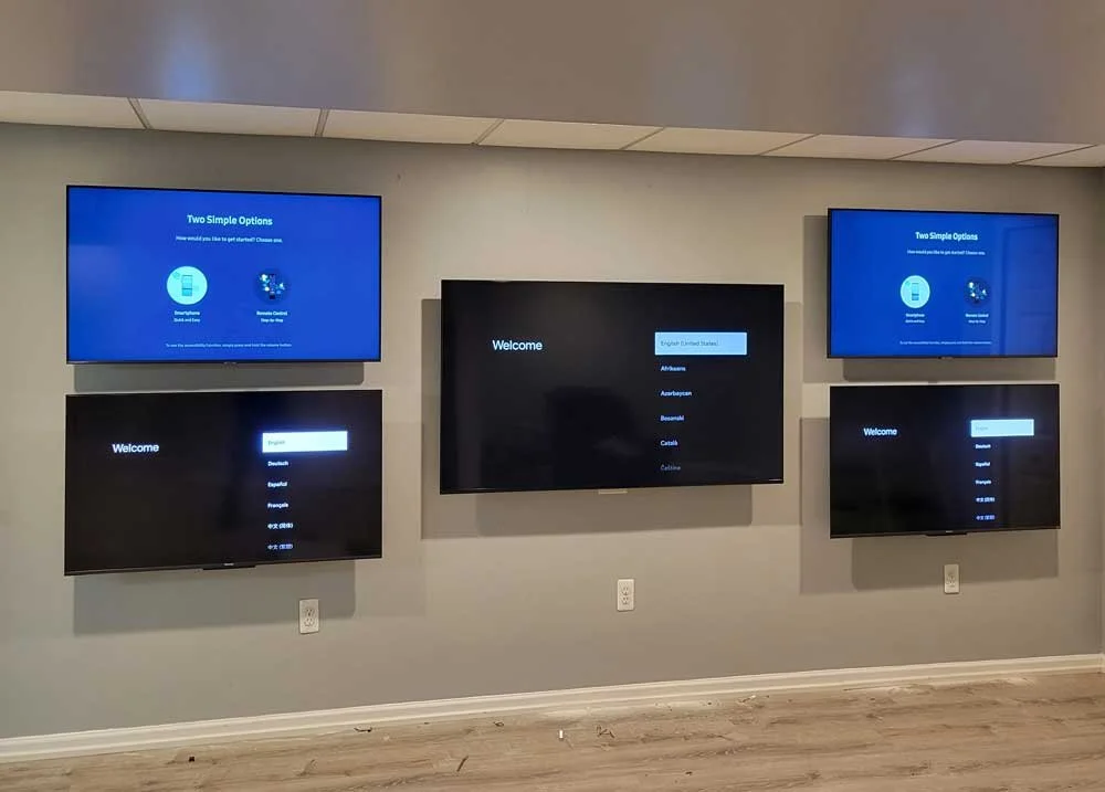 Five screens mounted on a gray wall in an indoor setting, displaying language options for a device setup or selection process.