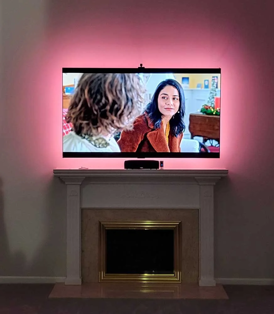 A television mounted above a fireplace shows two women talking, one with curly hair and the other with black hair, wearing a brown coat, in a cozy, decorated living room. The room is illuminated with pink ambient light.
