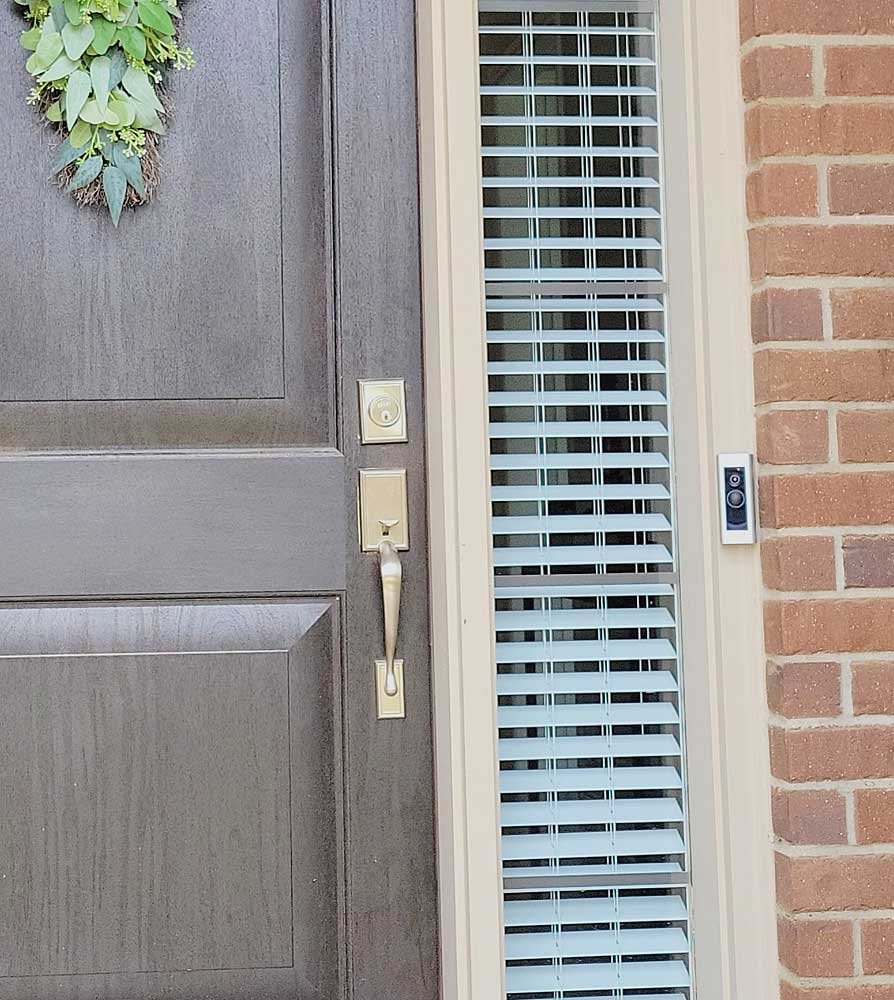 Front door with wreath, doorbell, and a door camera beside a window with blinds and brick wall.