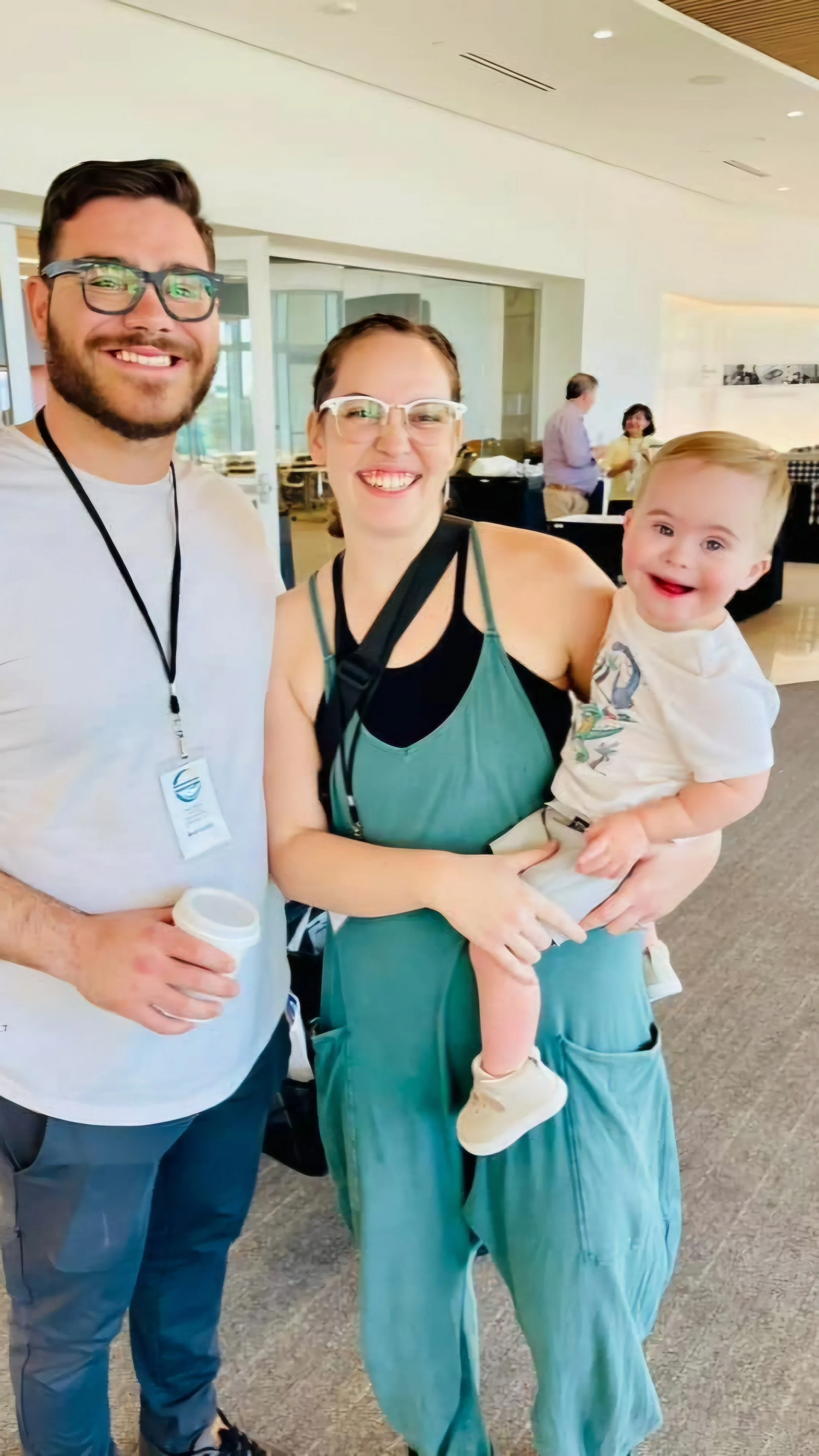 Smiling family in a bright, modern indoor space, with a man, woman, and young boy with Down syndrome, all wearing casual clothing.