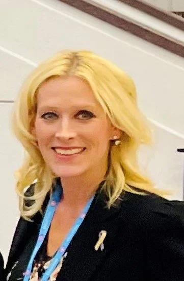 Portrait of a woman with blonde hair smiling, wearing a black blazer and a blue lanyard.