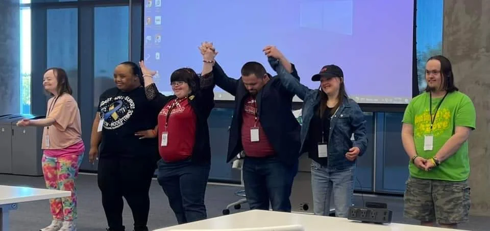Six people standing in a row, holding hands and raising them in celebration, inside a conference room with a large screen behind them.
