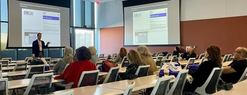 A presenter giving a lecture in a conference room with attendees seated at tables, two large screens displaying presentation slides, and modern office decor.